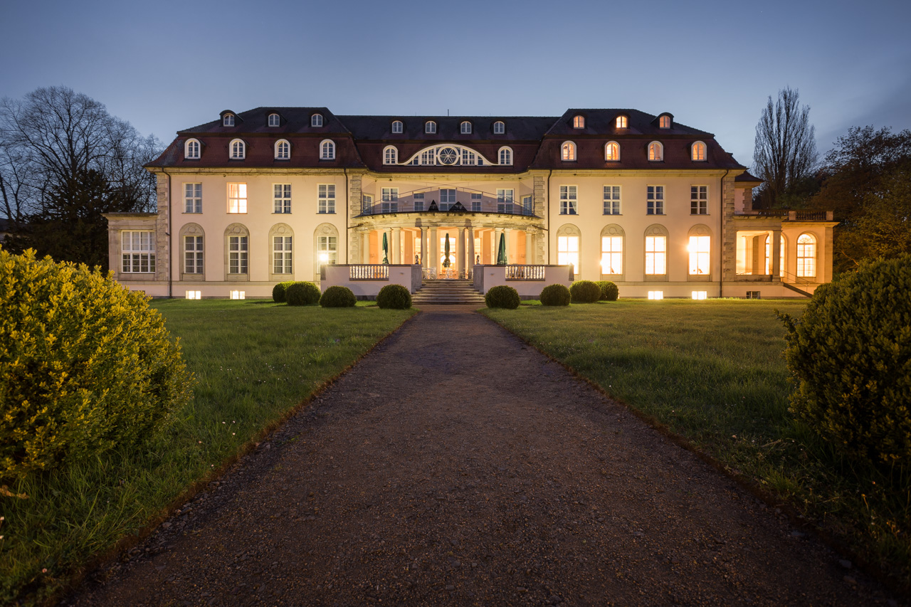Parkseite des Hotel Schloss Storkau am Abend mit beleuchteten Fenstern, Säulenportikus und geradem Kiesweg durch den Rasen