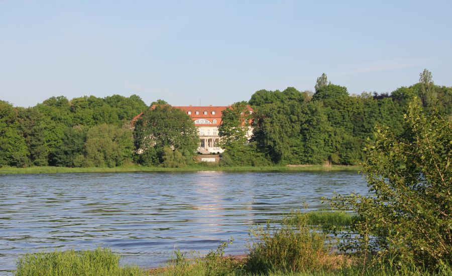 Schloss Storkau an der Elbe Blick über die Elbe auf das Hotel Schloss Storkau, das mit rotem Dach zwischen dichten Bäumen am bewaldeten Ufer liegt
