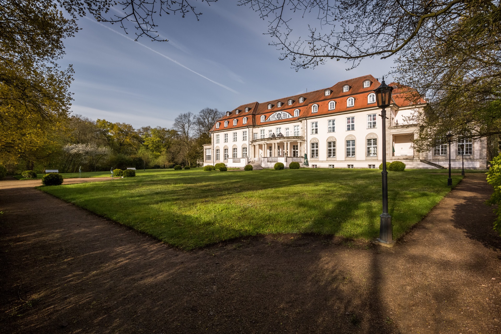 Parkseite des Hotel Schloss Storkau bei Sonnenschein mit weiter Rasenfläche, Parkwegen und historischer Laterne