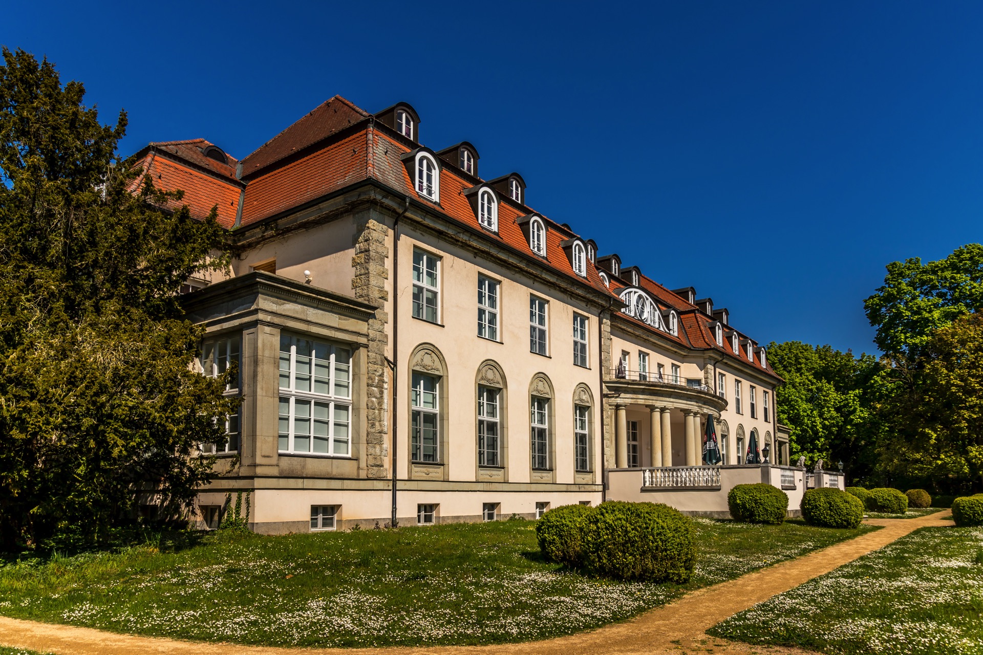 Seitenansicht des Hotel Schloss Storkau unter tiefblauem Himmel mit rotem Ziegeldach, Rundbogenfenstern und blühender Wiese