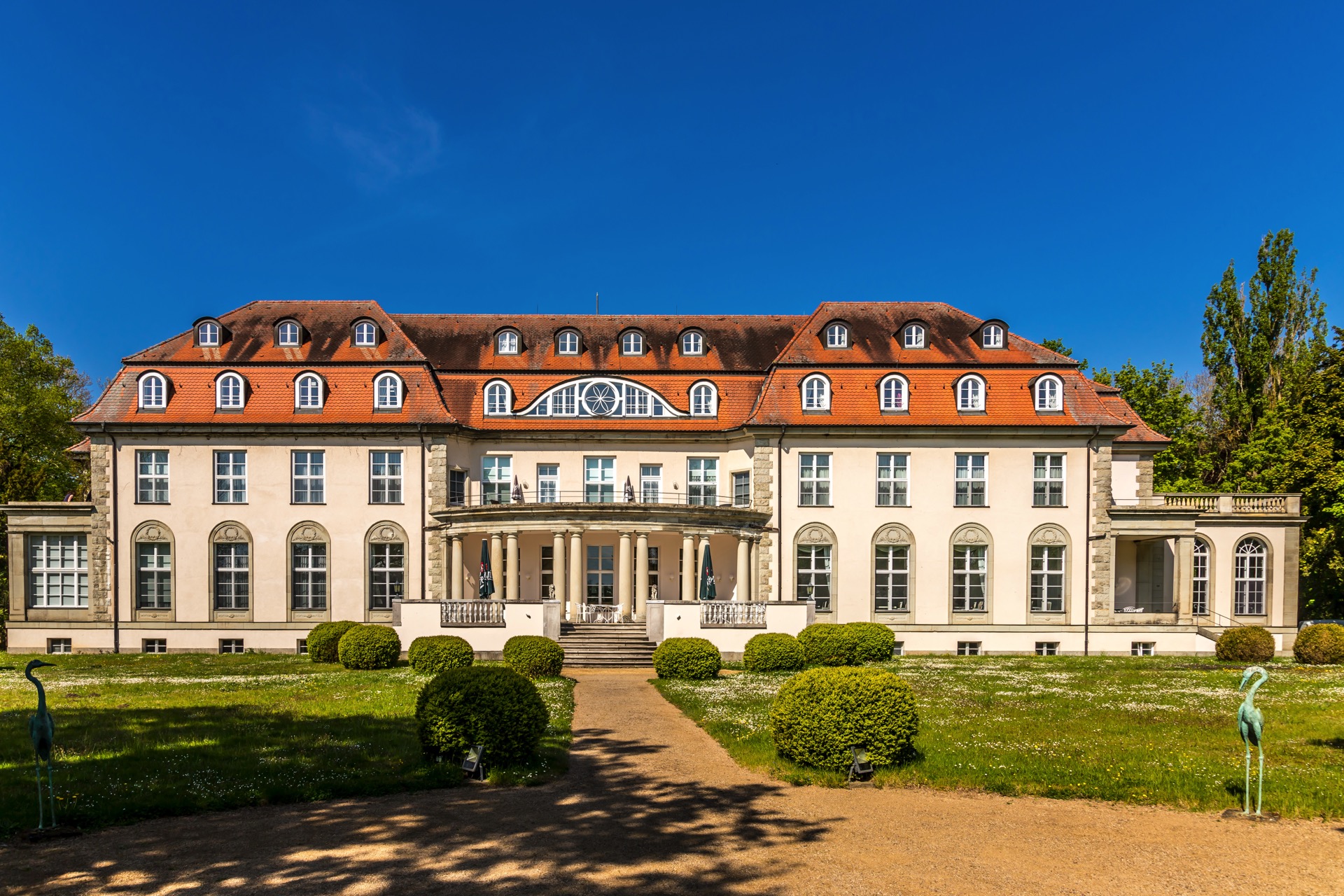 Gartenfassade des Hotel Schloss Storkau bei Sonnenschein mit rundem Säulenportikus, Freitreppe, Buchskugeln und Kranich-Skulpturen