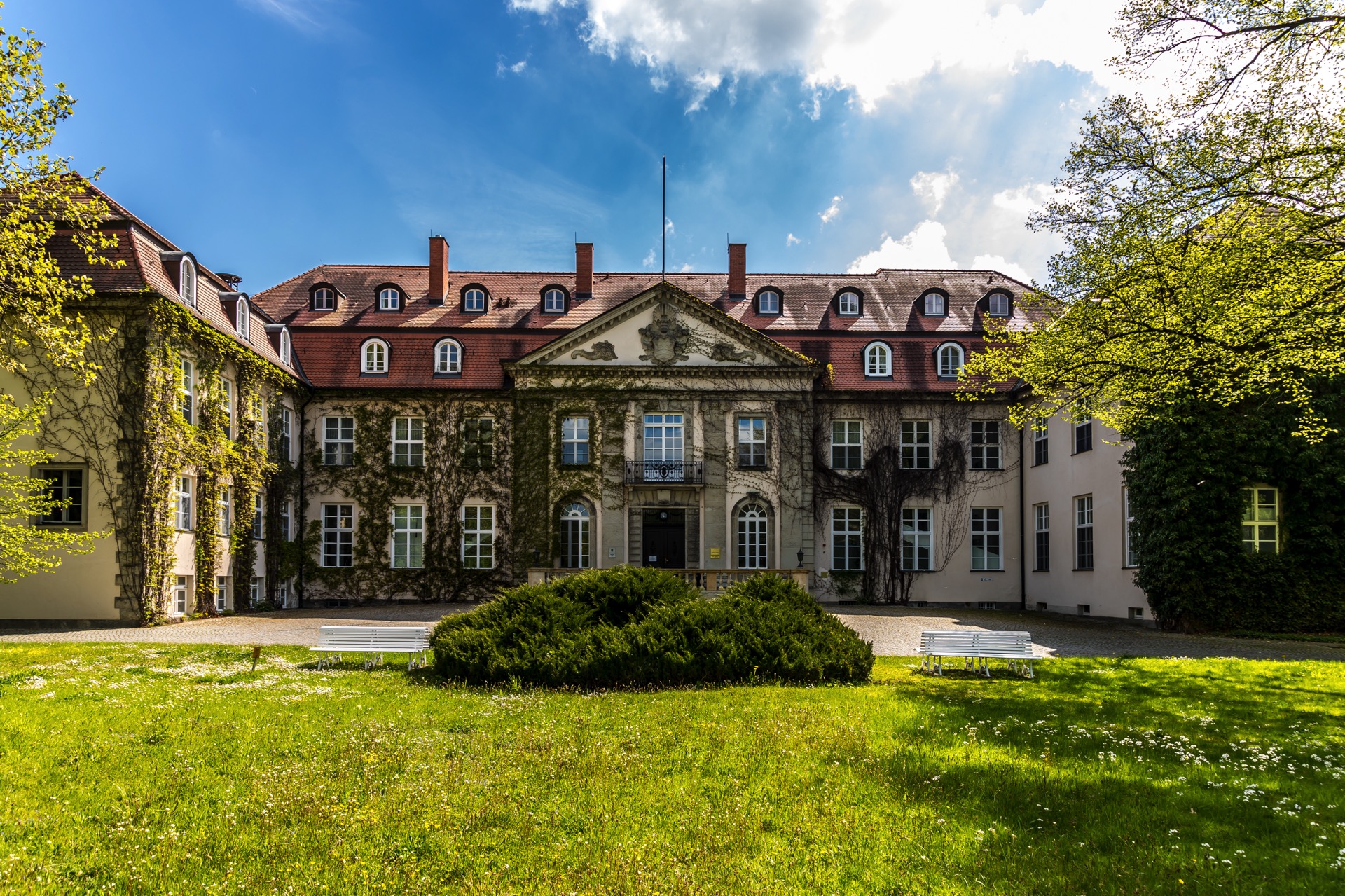 Hofseite des Hotel Schloss Storkau bei Sonnenschein mit efeubewachsener Fassade, Wappengiebel und weißen Gartenbänken auf dem Rasenrondell