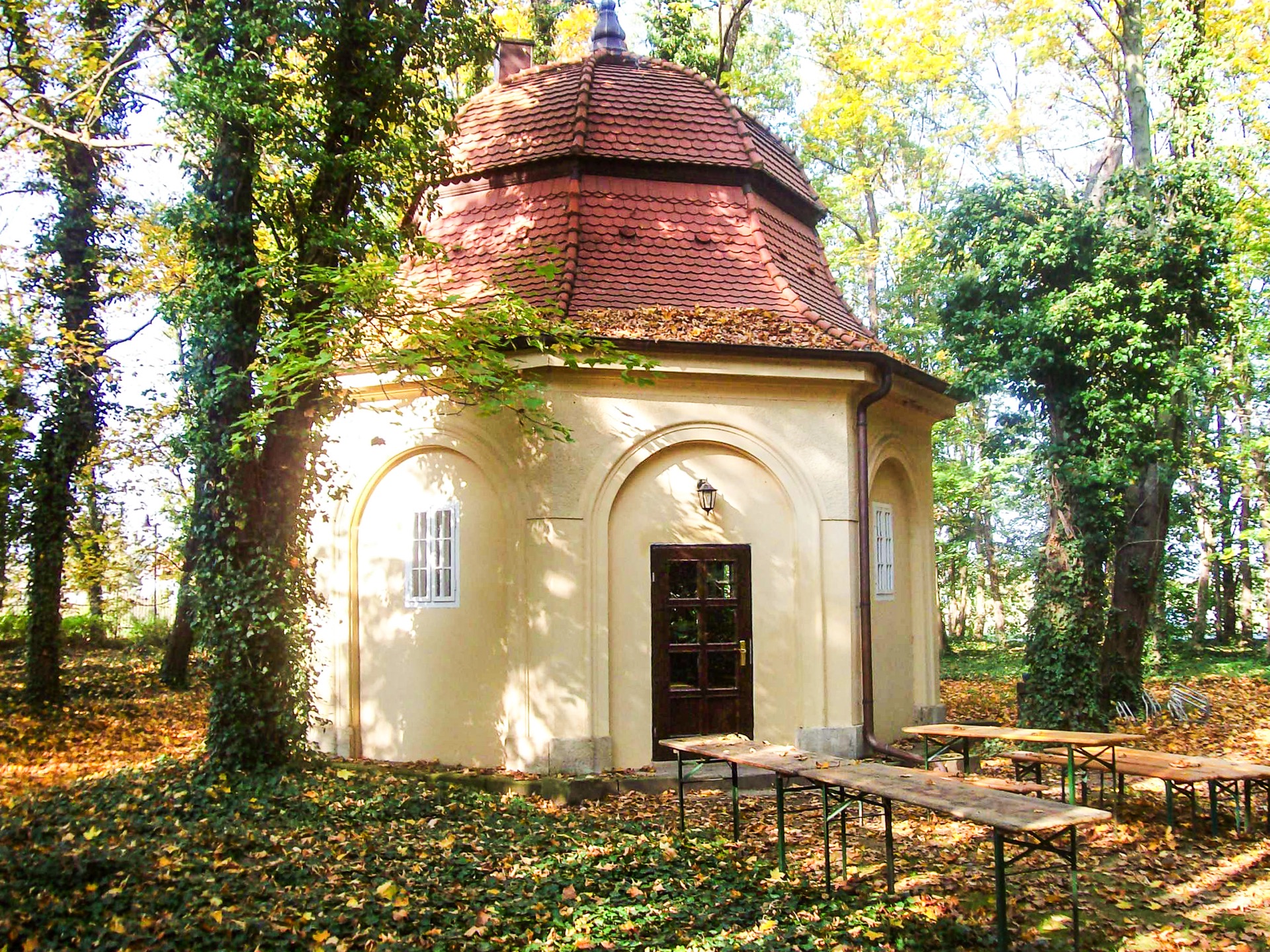 Achteckiges Brunnenhaus im Schlosspark des Hotel Schloss Storkau mit geschwungenem Ziegeldach, Rundbogennischen und Bierzeltgarnituren im Herbstlaub