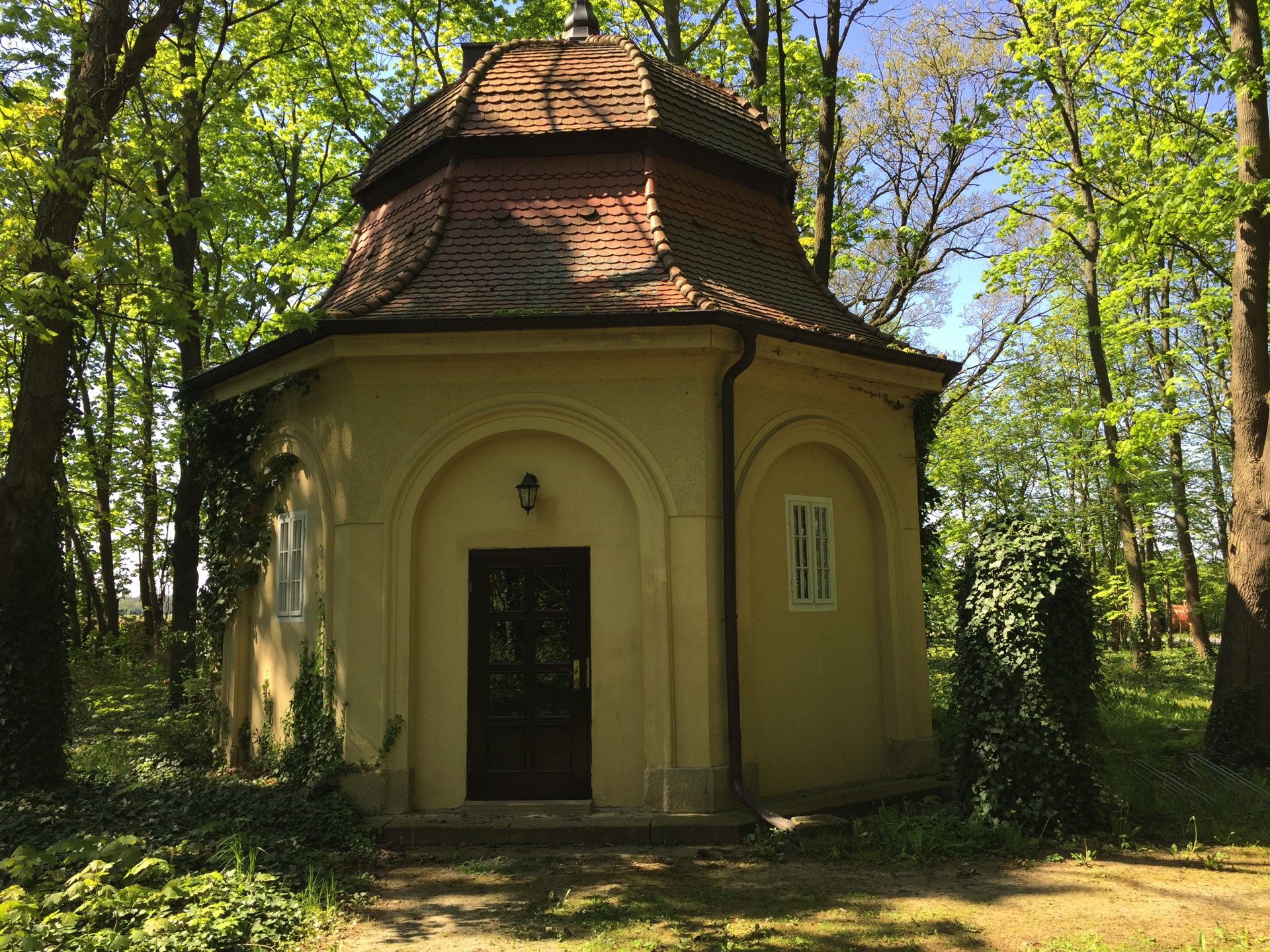 Brunnenhaus im Schlosspark des Hotel Schloss Storkau, achteckiger Pavillon mit rotem Schindeldach und dunkler Holztür zwischen frühlingsgrünen Bäumen