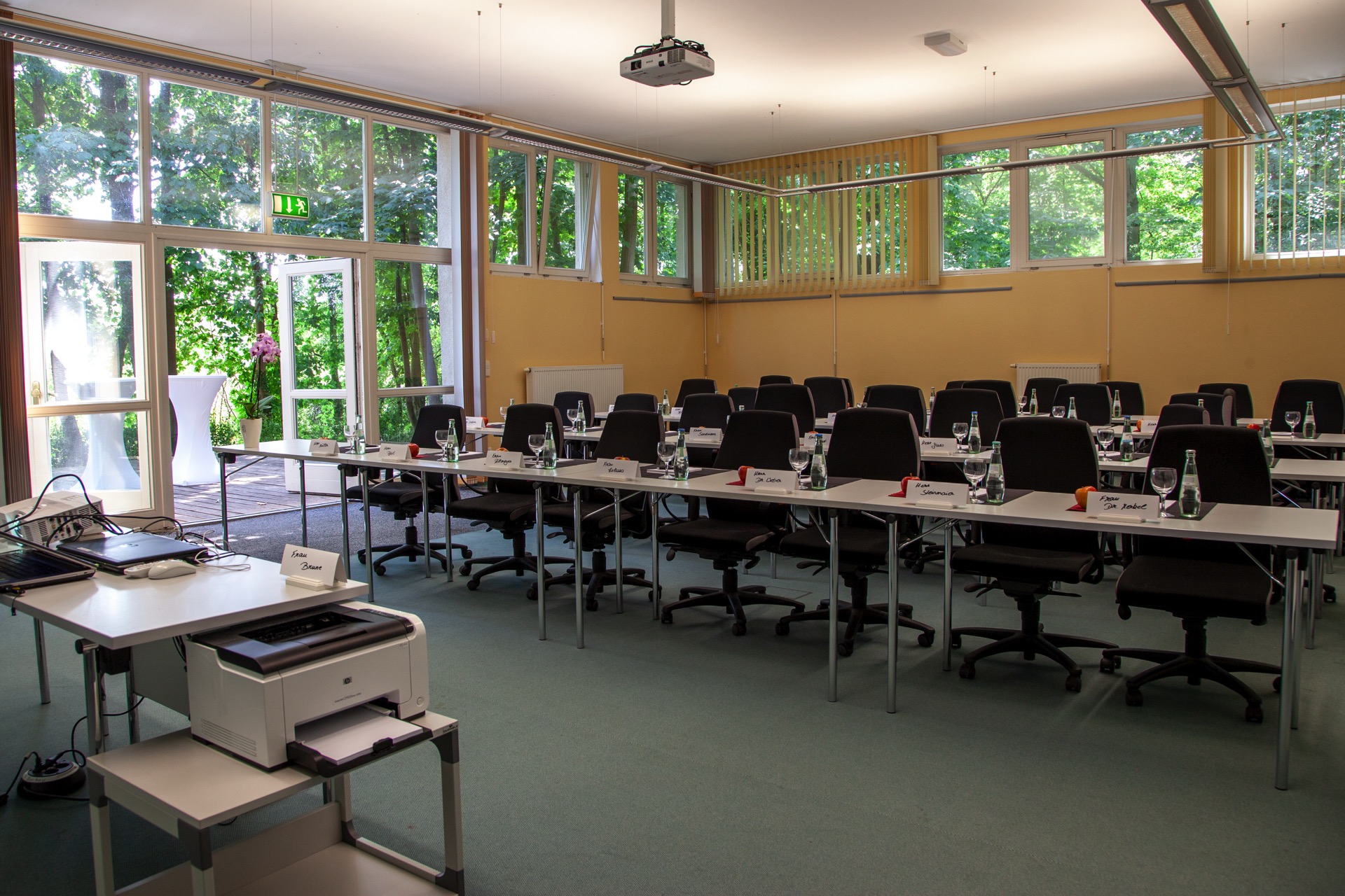 Konferenzraum mit Paneltisch, schwarzen Stühlen, Namensplaketten, Wassergläsern und Äpfeln, im Hintergrund große Fenster mit Blick ins Grüne.