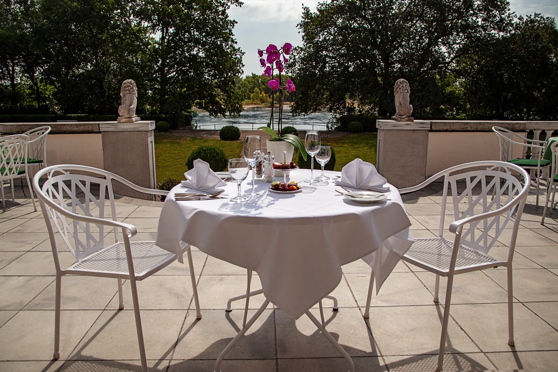 Runder Tisch mit weißem Tischtuch und Orchidee auf der Elbterrasse des Hotel Schloss Storkau, dahinter Löwenfiguren und die Elbe