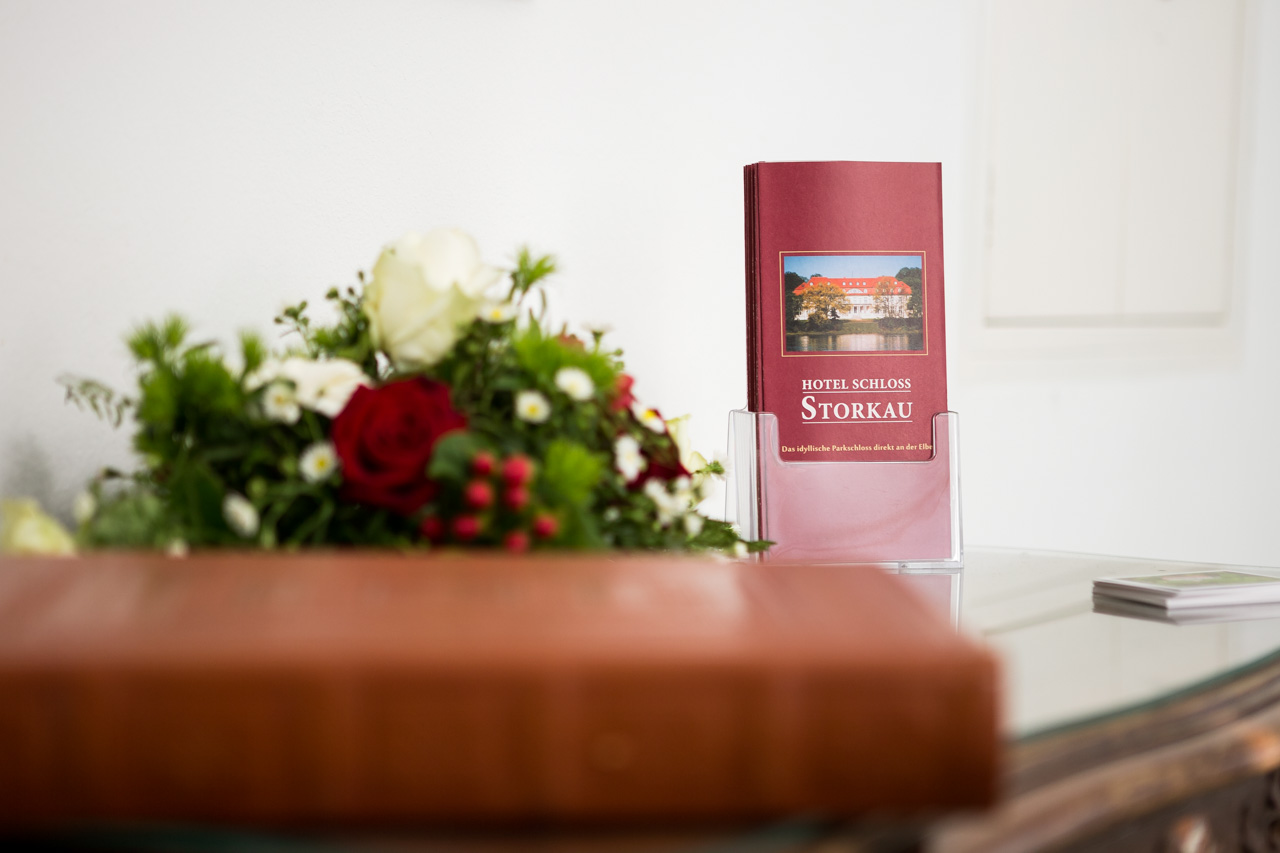 Hausprospekt des Hotel Schloss Storkau in einem Acrylständer, davor ein Blumengesteck mit Rosen