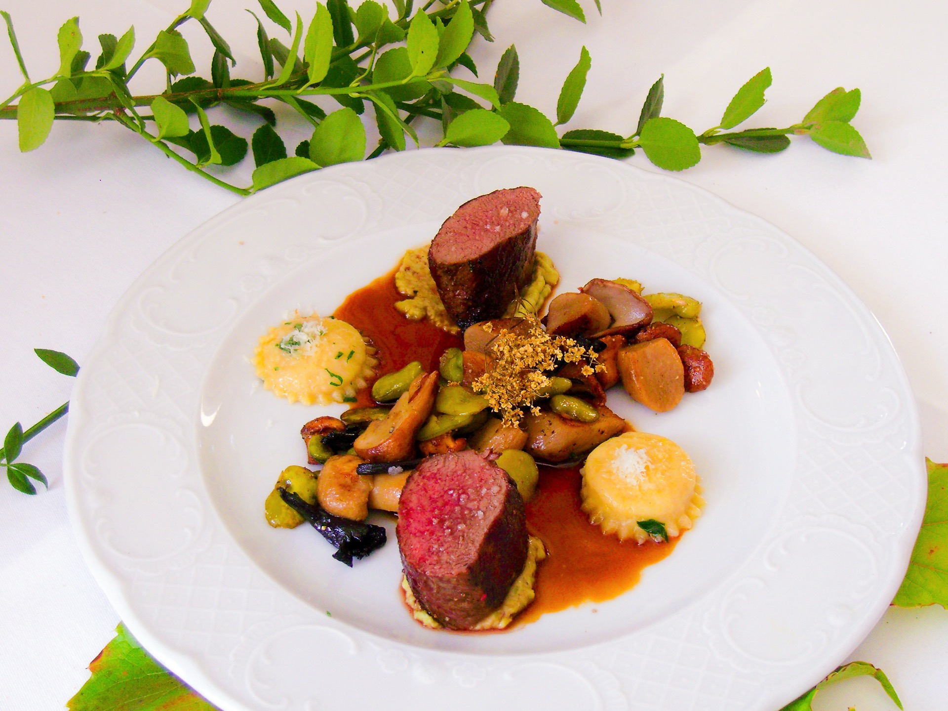 Rosa gebratene Fleischmedaillons mit Ravioli, Pilzen und dicken Bohnen in dunkler Jus auf weißem Teller im Hotel Schloss Storkau