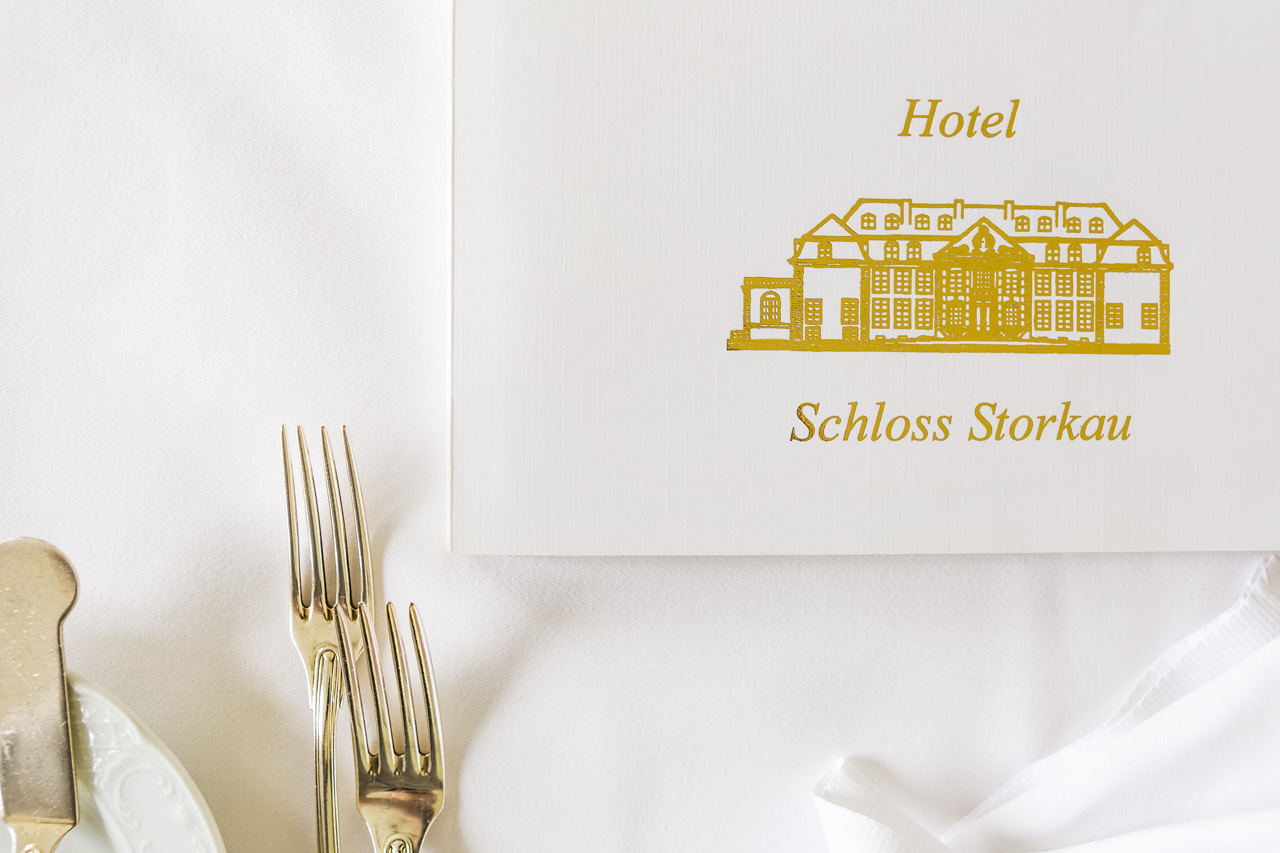 Speisekarte mit Goldprägung Speisekarte mit goldgeprägtem Schriftzug und Schlossfassade des Hotel Schloss Storkau neben Silberbesteck