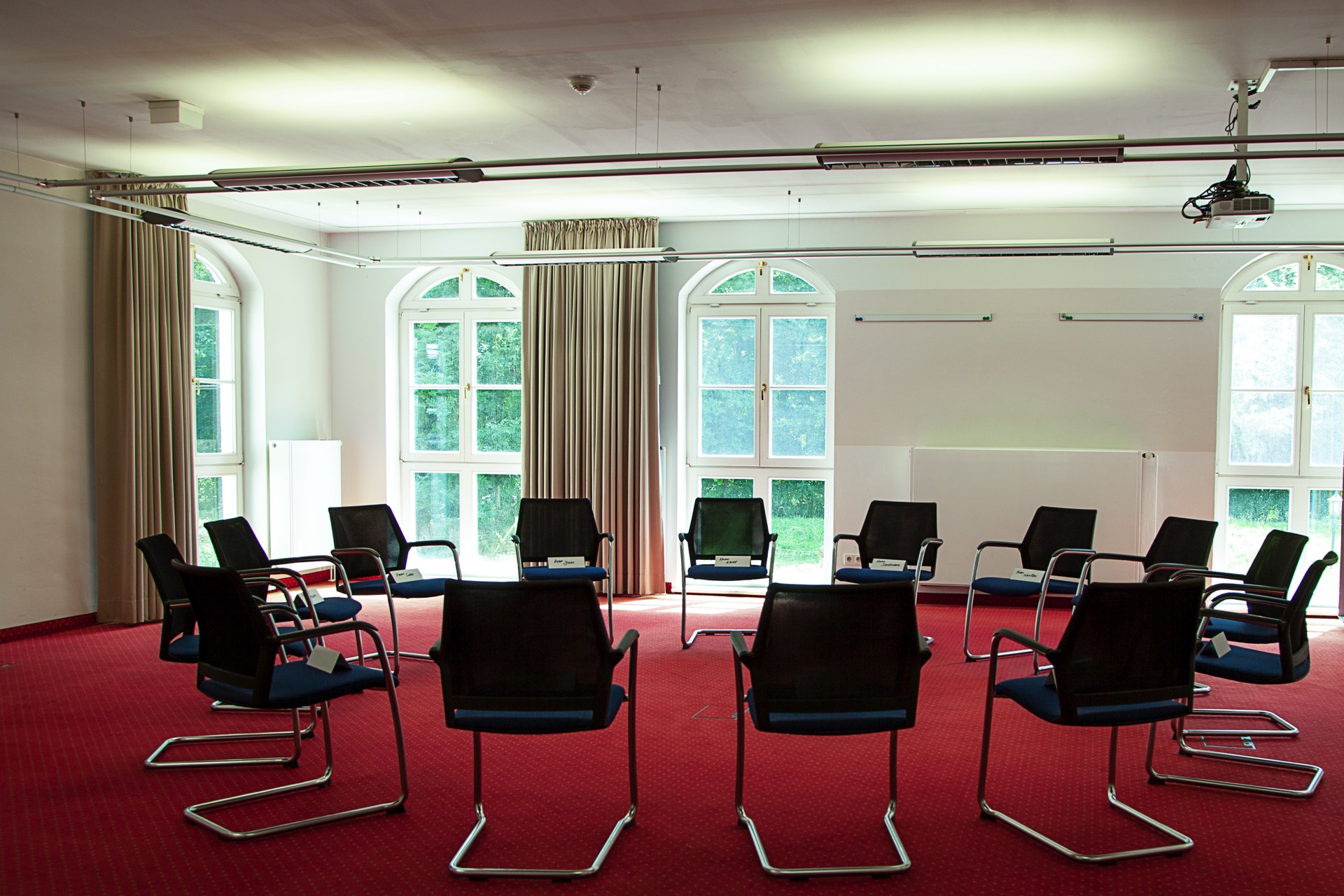 Großer Konferenzraum mit schwarzen Designstühlen auf rotem Teppich, großen Fenstern und Vorhängen vor grüner Parklandschaft.