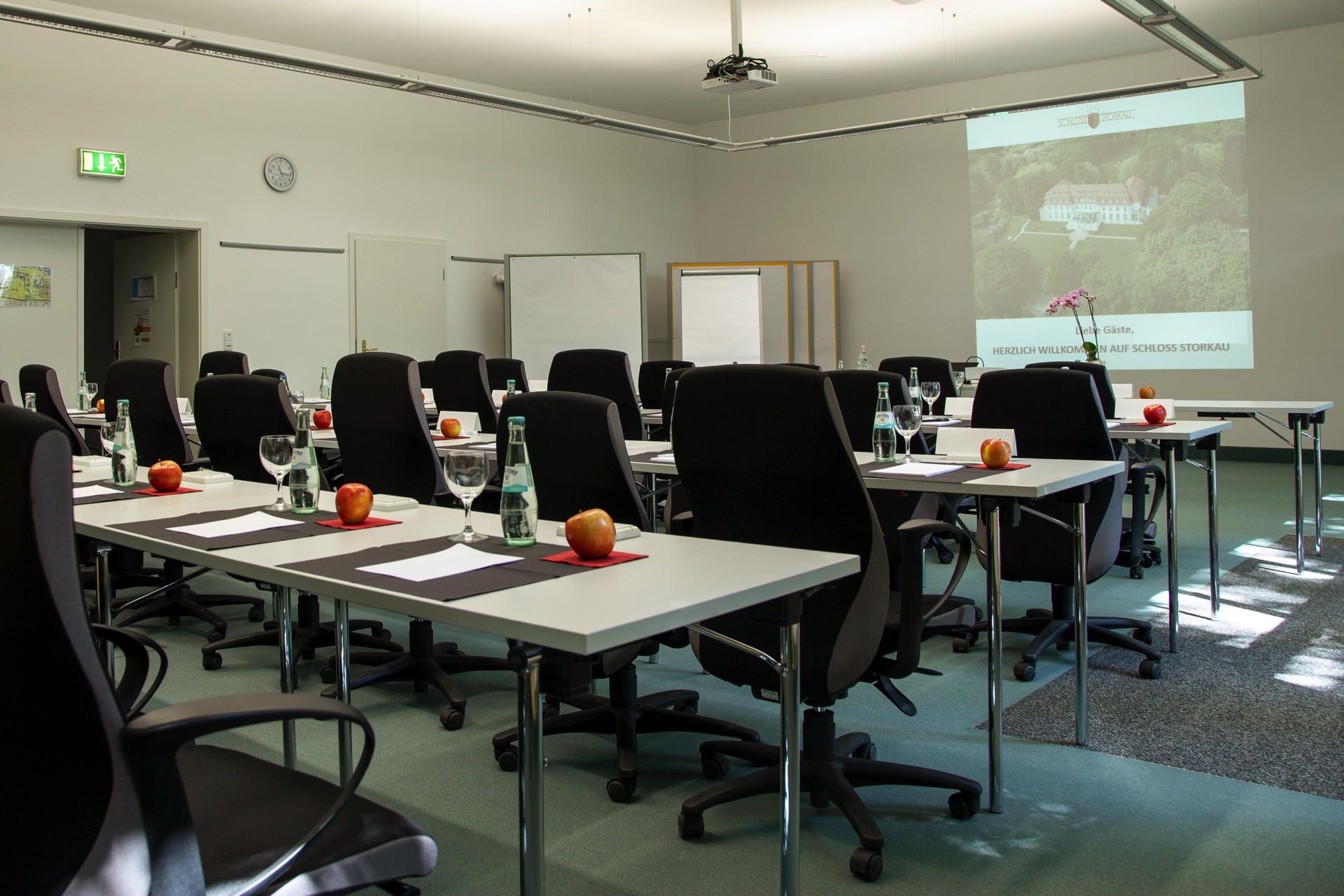 Tagungsraum im Kavaliershaus mit langen Konferenztischen, schwarzen Bürostühlen, frischen Äpfeln und Beamer-Projektion der Schloss-Storkau-Website.
