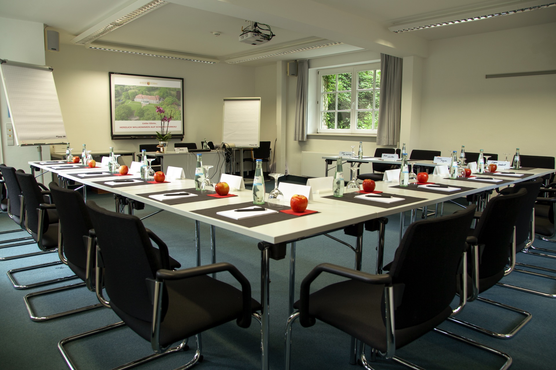 Tagungsraum im Kavaliershaus mit langen Konferenztischen, schwarzen Bürostühlen, frischen Äpfeln und Beamer-Projektion der Schloss-Storkau-Website.