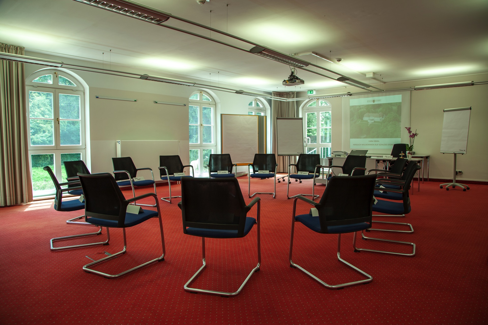 Großer Konferenzraum mit schwarzen Designstühlen auf rotem Teppich, großen Fenstern und Vorhängen vor grüner Parklandschaft.