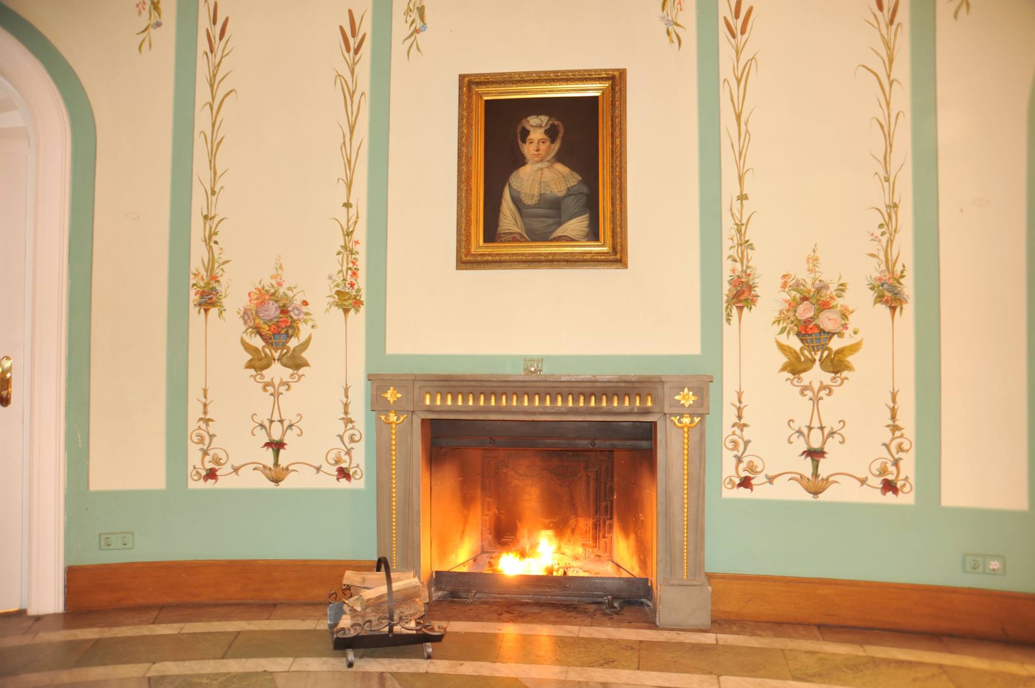 Brennender Kamin im Salon des Hotel Schloss Storkau mit Damenporträt in Goldrahmen und mintgrünen Wandmalereien mit Blumenmotiven