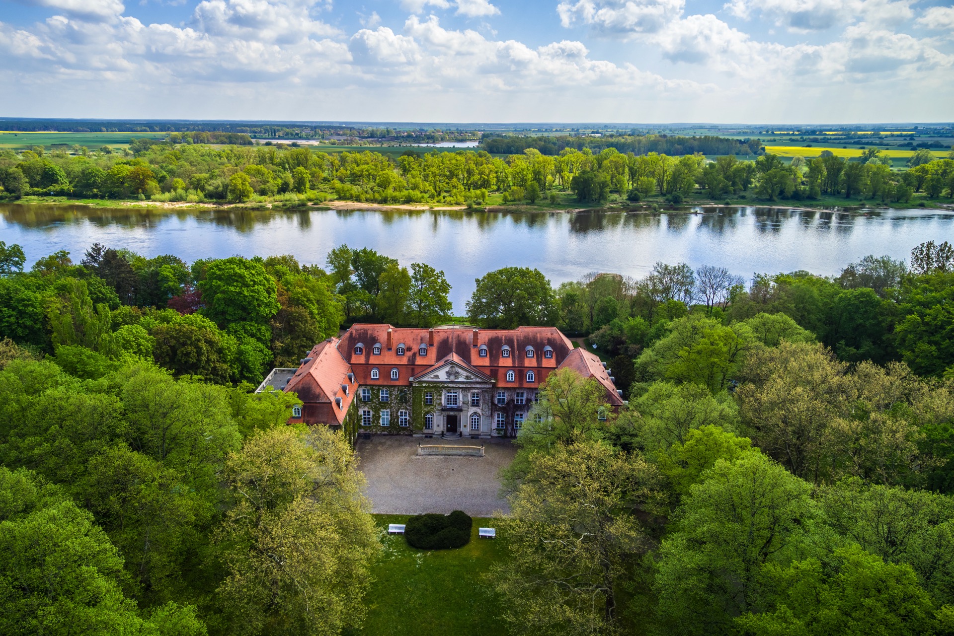 Drohnenaufnahme des Hotel Schloss Storkau mit efeubewachsener Hoffassade, davor der Schlosshof, dahinter die breite Elbe und die Flusslandschaft