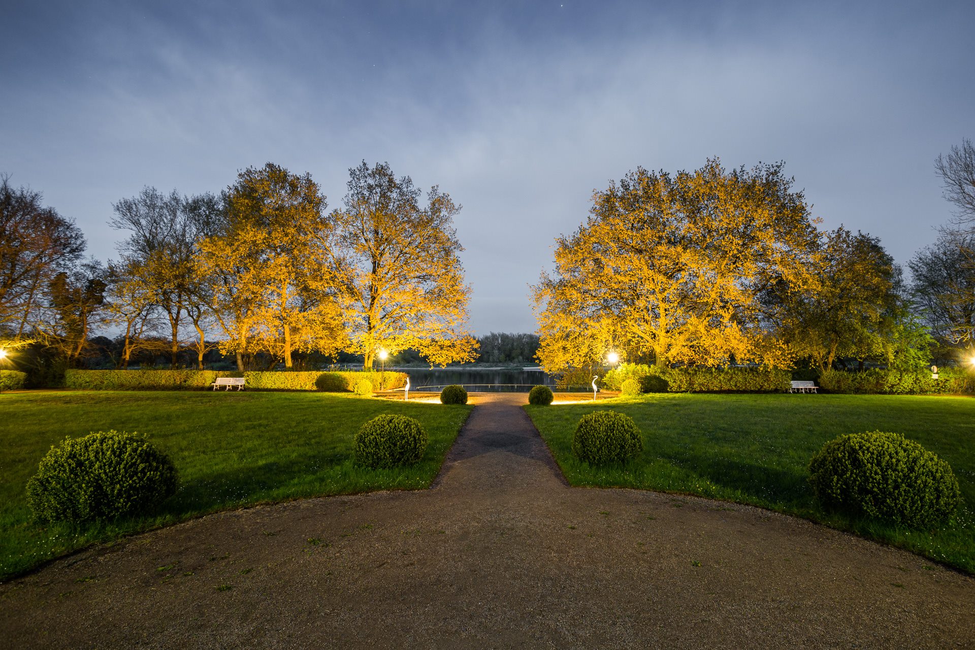 Nächtlich angestrahlte Bäume im Schlosspark des Hotel Schloss Storkau mit Wegeachse zum Elbufer, weißen Parkbänken und Reiherfiguren