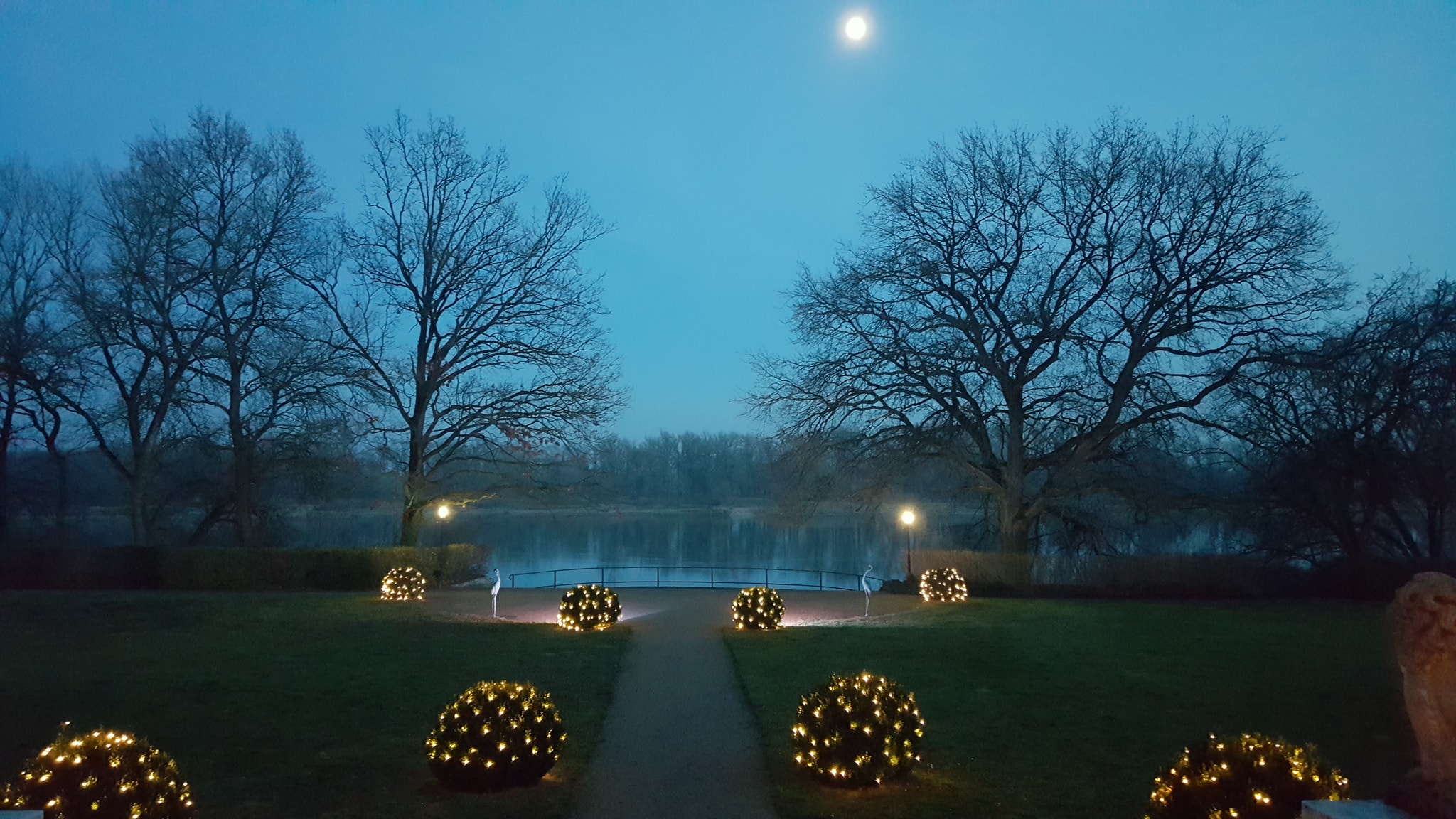 Schlosspark des Hotel Schloss Storkau in der Dämmerung mit Mond, beleuchteten Buchskugeln und Blick über die Brüstung zur Elbe
