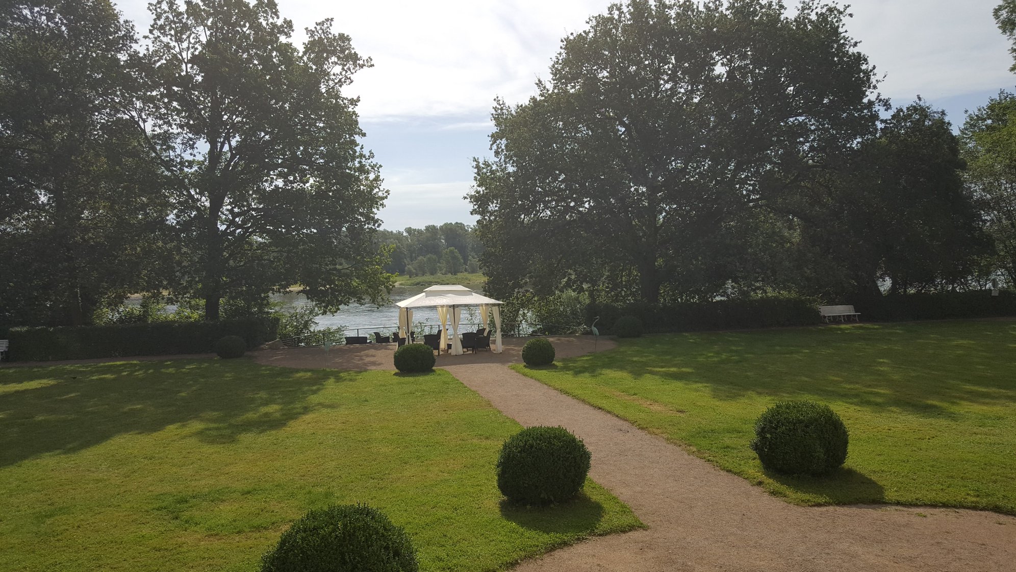 Weißer Gartenpavillon mit Sitzgruppe auf der Uferterrasse im Schlosspark des Hotel Schloss Storkau, dahinter die Elbe
