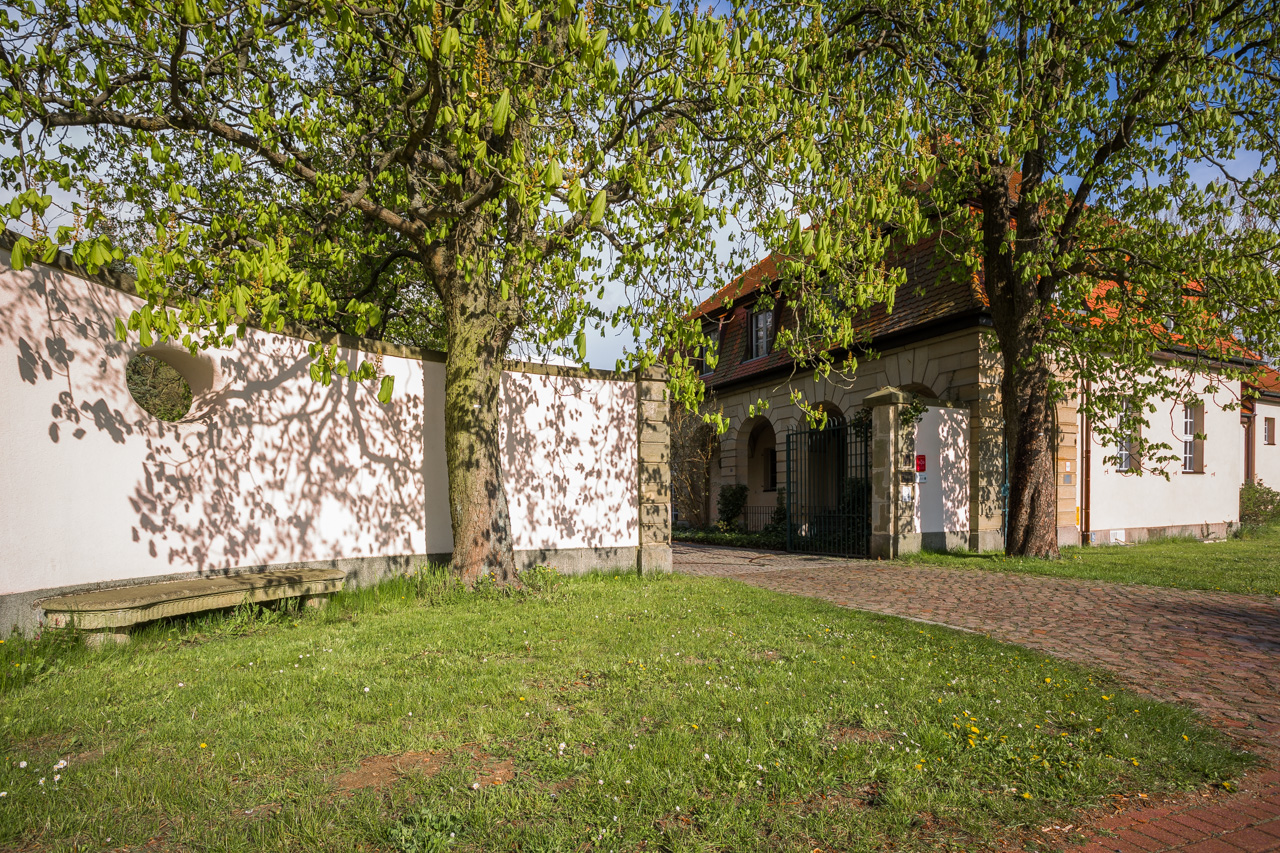 Einfahrt am Pförtnerhaus des Hotel Schloss Storkau mit weißer Gartenmauer, rundem Mauerfenster, Steinbank und gepflastertem Weg unter Kastanien