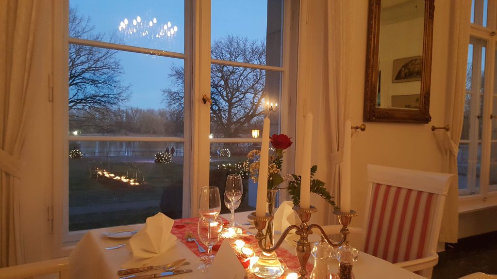 Romantisch gedeckter Fenstertisch mit Kerzen und roter Rose im Hotel Schloss Storkau, davor der abendlich beleuchtete Schlosspark
