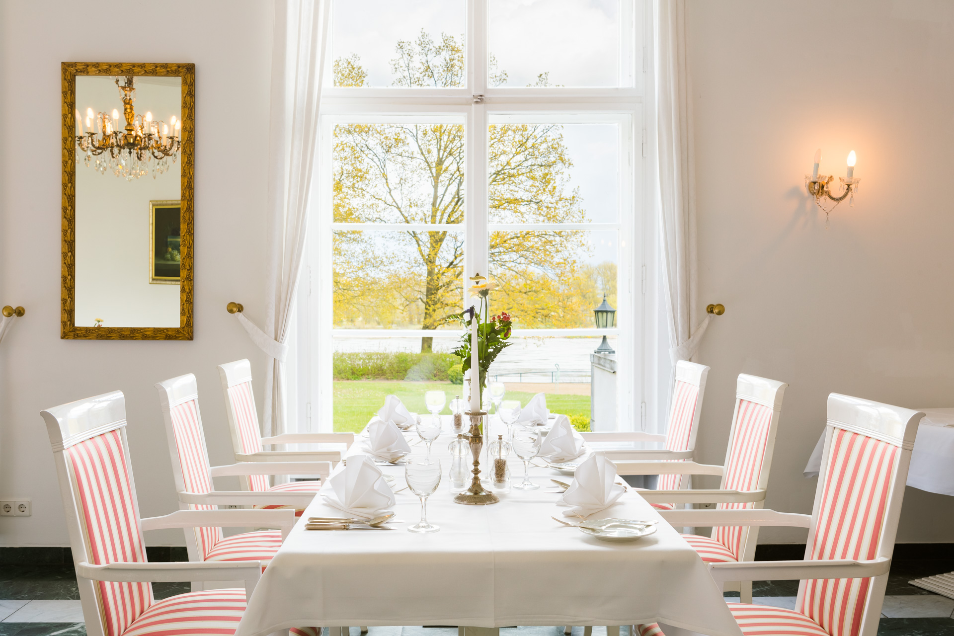 Lange Tafel mit rot-weiß gestreiften Stühlen am hohen Fenster im Restaurant des Hotel Schloss Storkau mit Blick in den Park