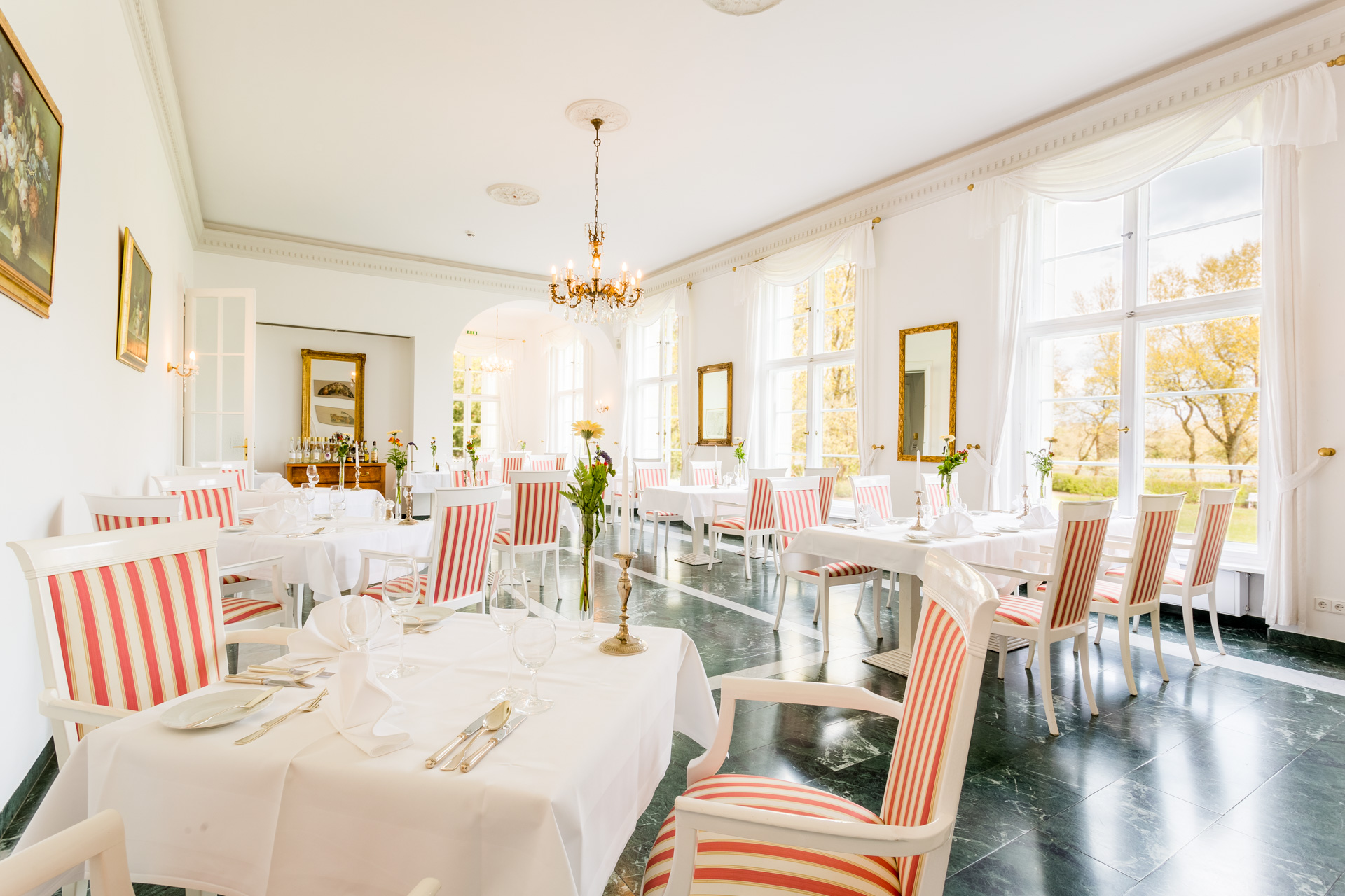 Lichtdurchfluteter Restaurantsaal im Hotel Schloss Storkau mit weiß gedeckten Tischen, Blumen und rot-weiß gestreiften Stühlen