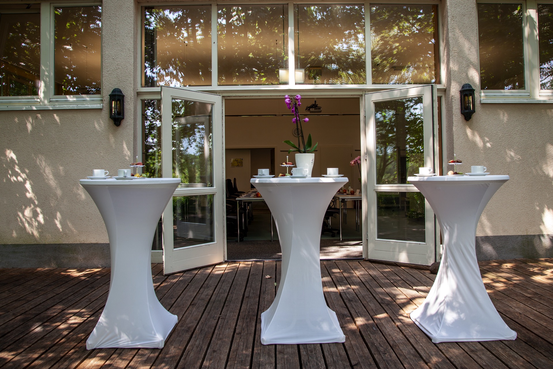 Stehtische auf der TerrasseKaffeepause auf der Terrasse vor dem Tagungsraum im Schloss Storkau. Drei weiß eingedeckte Stehtische mit Kaffeegedeck und Orchidee auf einer Holzterrasse vor der geöffneten Glasfront eines Tagungsraums.