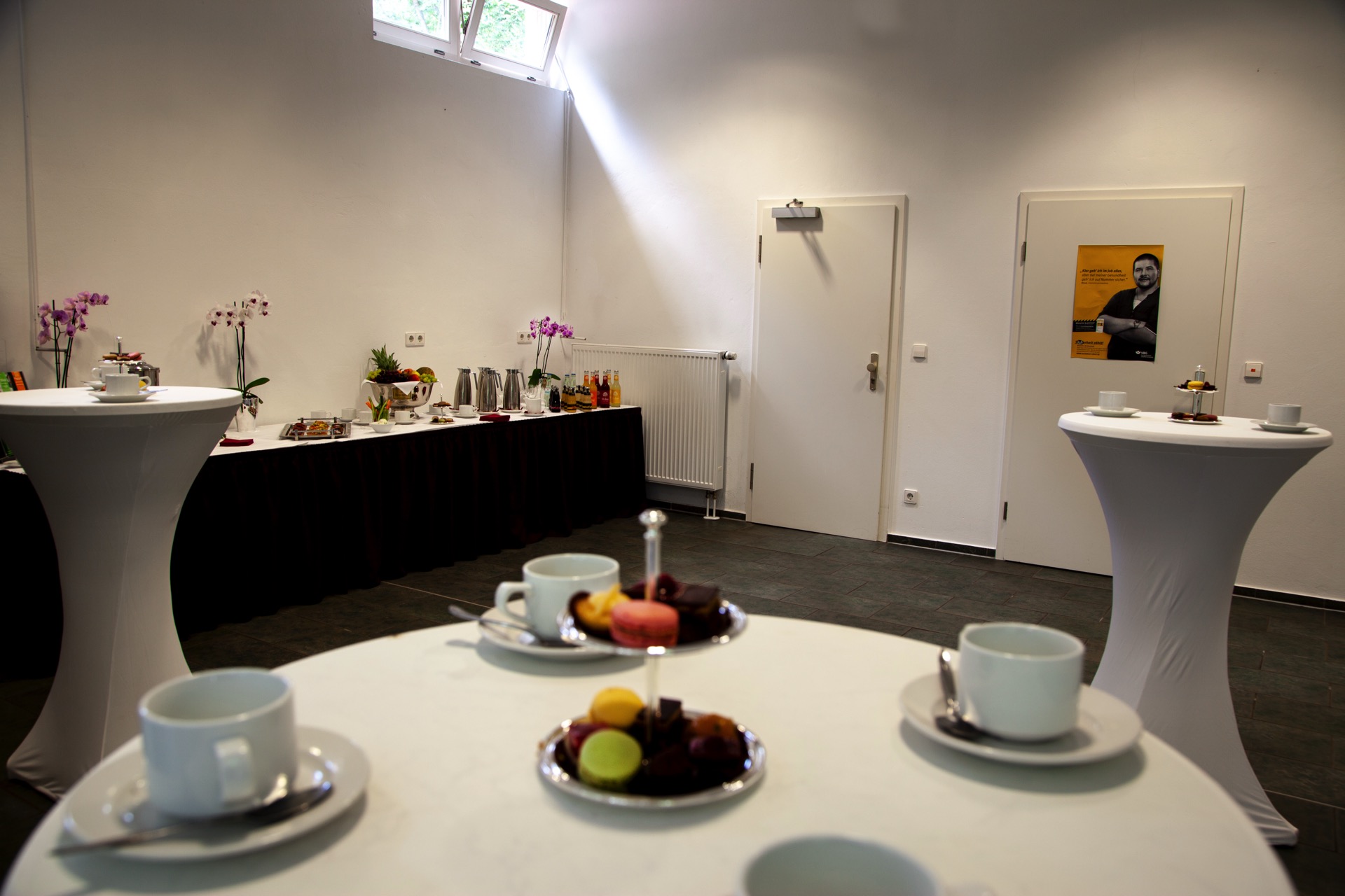 Tagungsraum mit weißen Stehtischen, Kaffeegedeck und Etageren mit Macarons sowie einem Buffet mit Obst, Getränken und Orchideen.