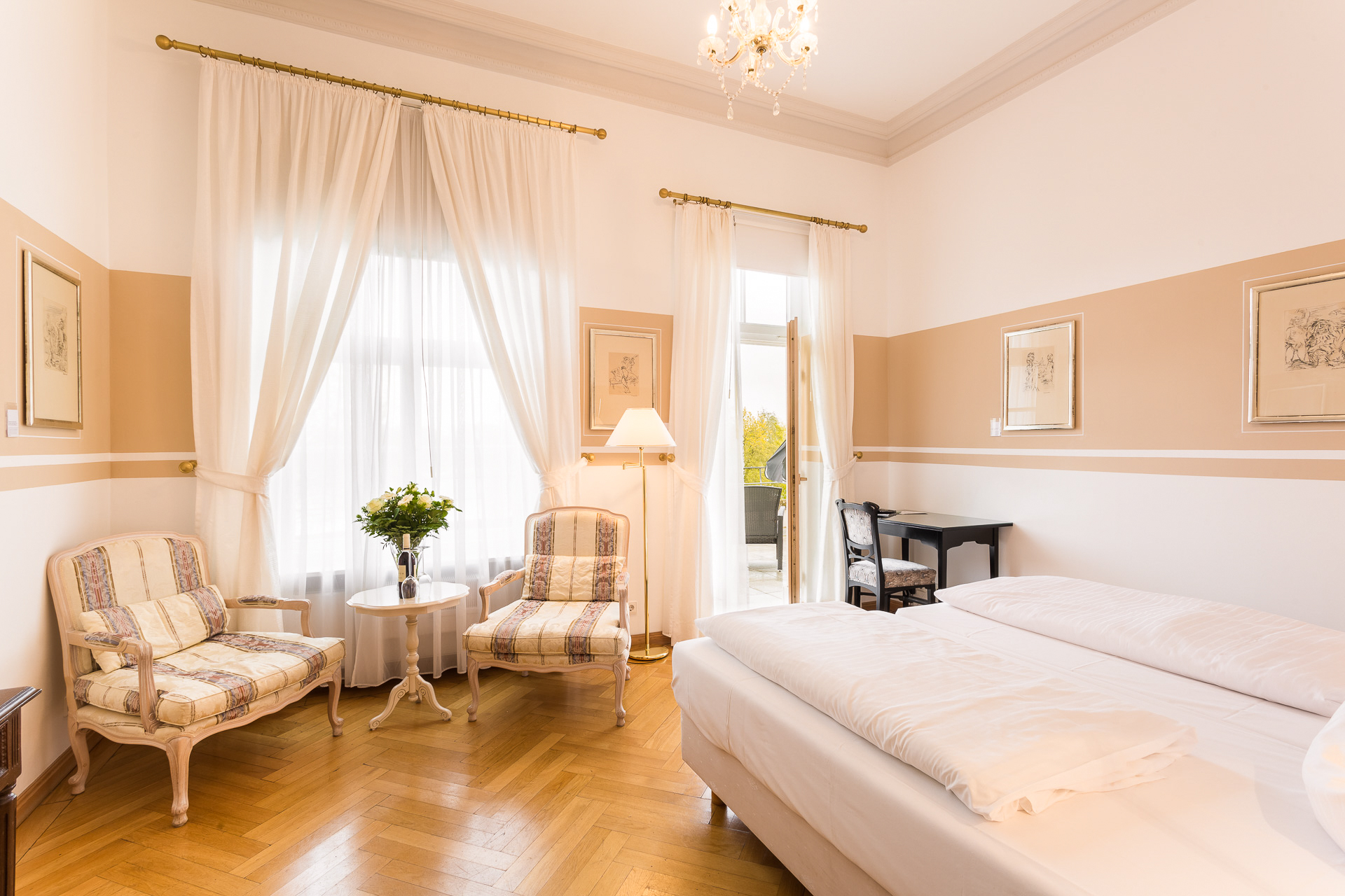 Deluxe-Zimmer im Hotel Schloss Storkau mit Doppelbett, Sitzecke mit gestreiften Sesseln und geöffneter Tür zur Terrasse