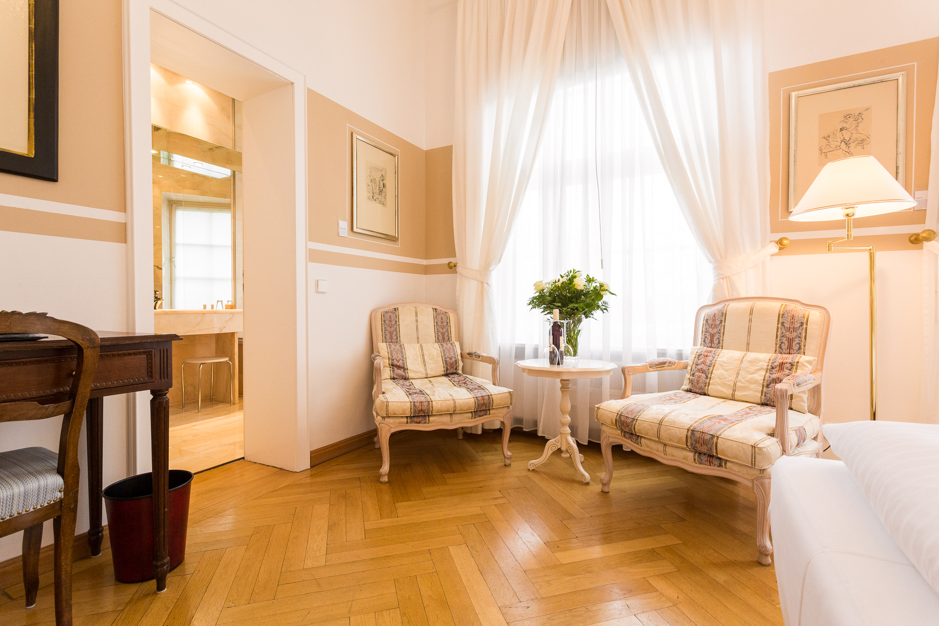 Sitzecke eines Deluxe-Zimmers im Hotel Schloss Storkau mit zwei gestreiften Sesseln, rundem Beistelltisch und Blick ins Marmorbad