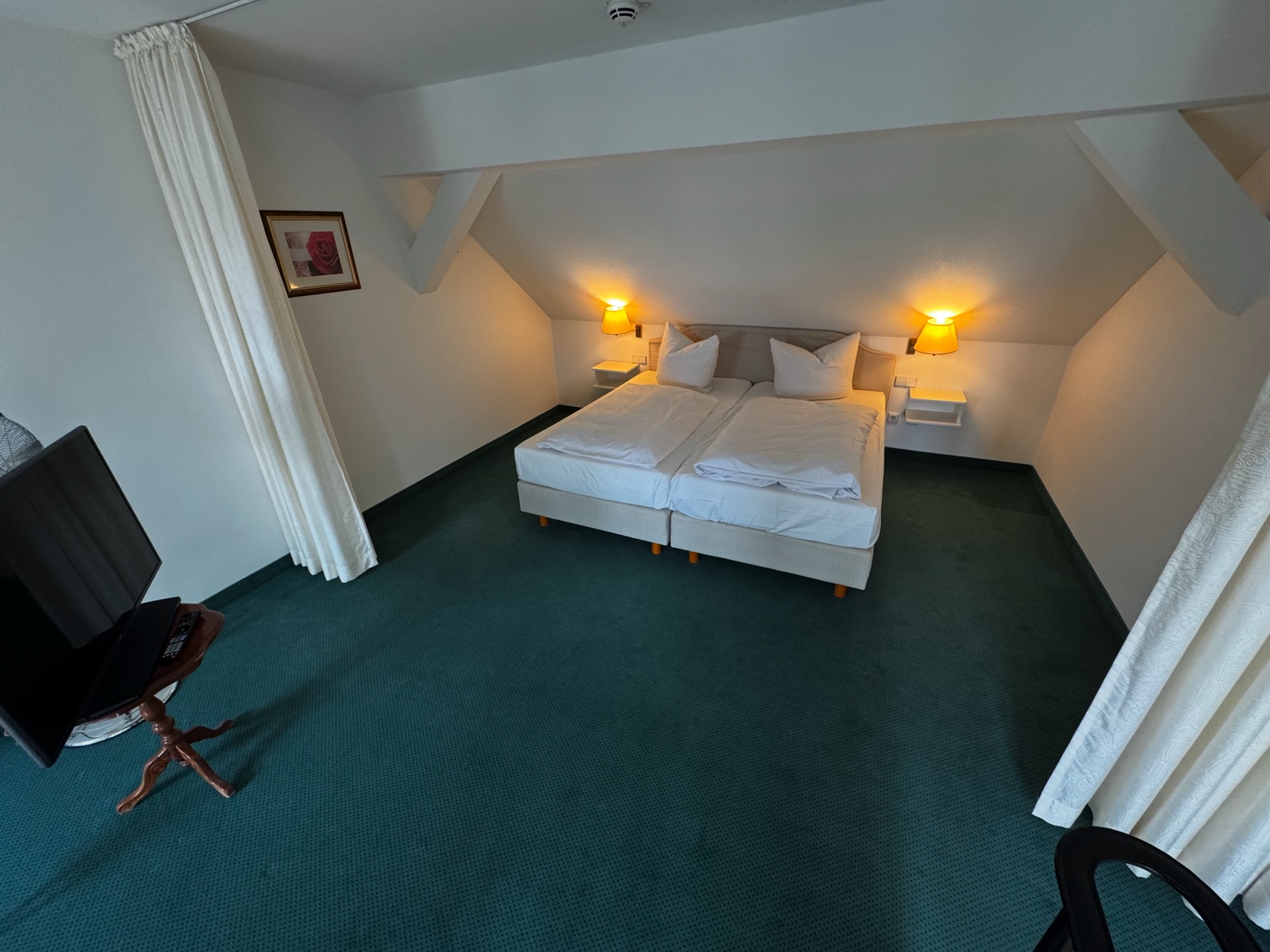 Schlafbereich eines Deluxe-Zimmers im Dachgeschoss des Hotel Schloss Storkau mit Doppelbett, grünem Teppichboden und Vorhängen als Raumteiler