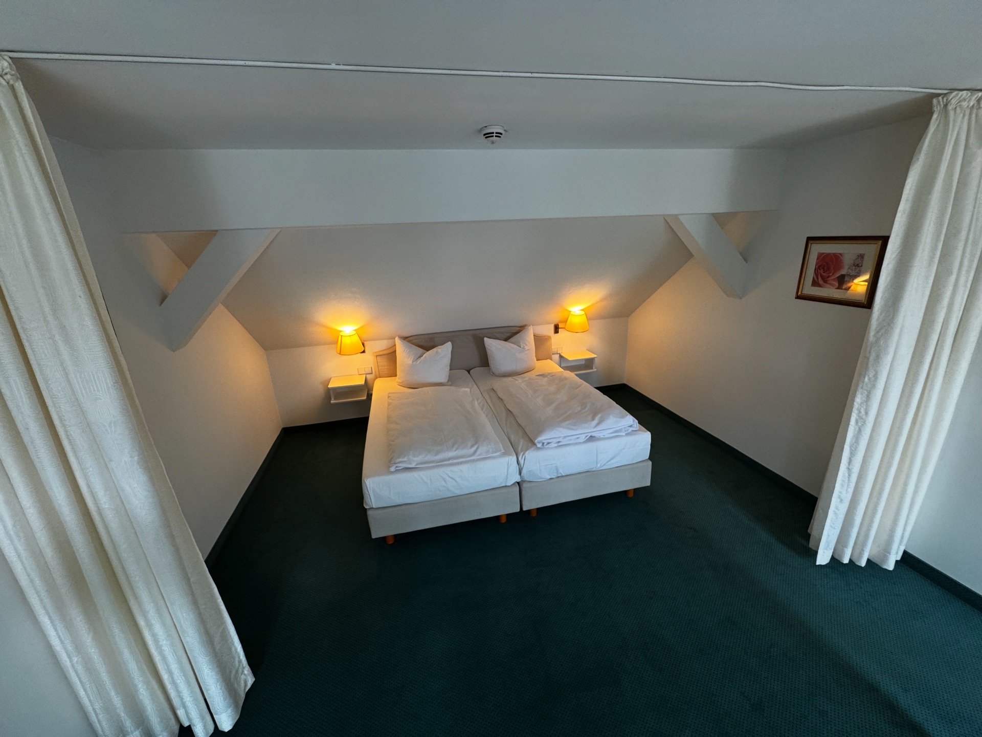 Doppelbett unter der Dachschräge in einem Deluxe-Zimmer im Hotel Schloss Storkau mit beleuchteten Wandlampen und grünem Teppichboden