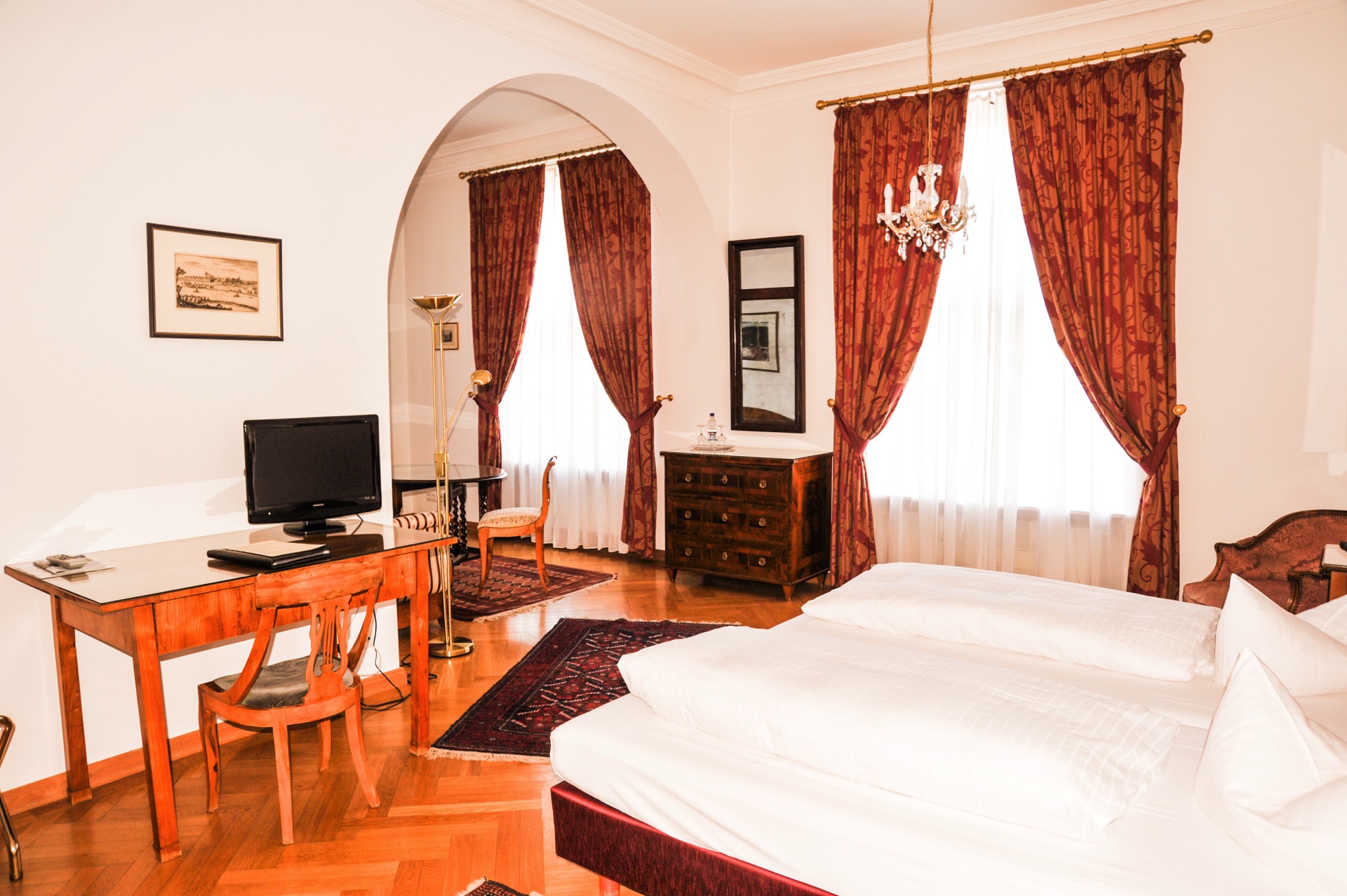 Deluxe-Zimmer im Hotel Schloss Storkau mit rot gemusterten Vorhängen, Kronleuchter, antiker Kommode und Doppelbett