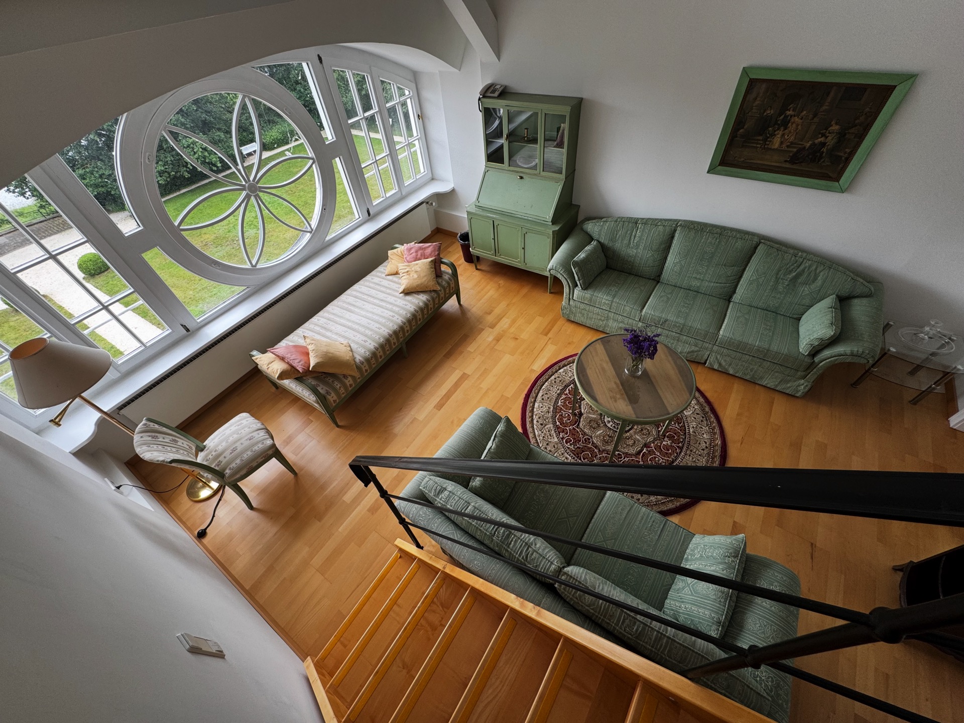 Wohnbereich einer Deluxe-Maisonette im Hotel Schloss Storkau mit grüner Polstergarnitur, Ornament-Rundfenster und Blick in den Schlosspark