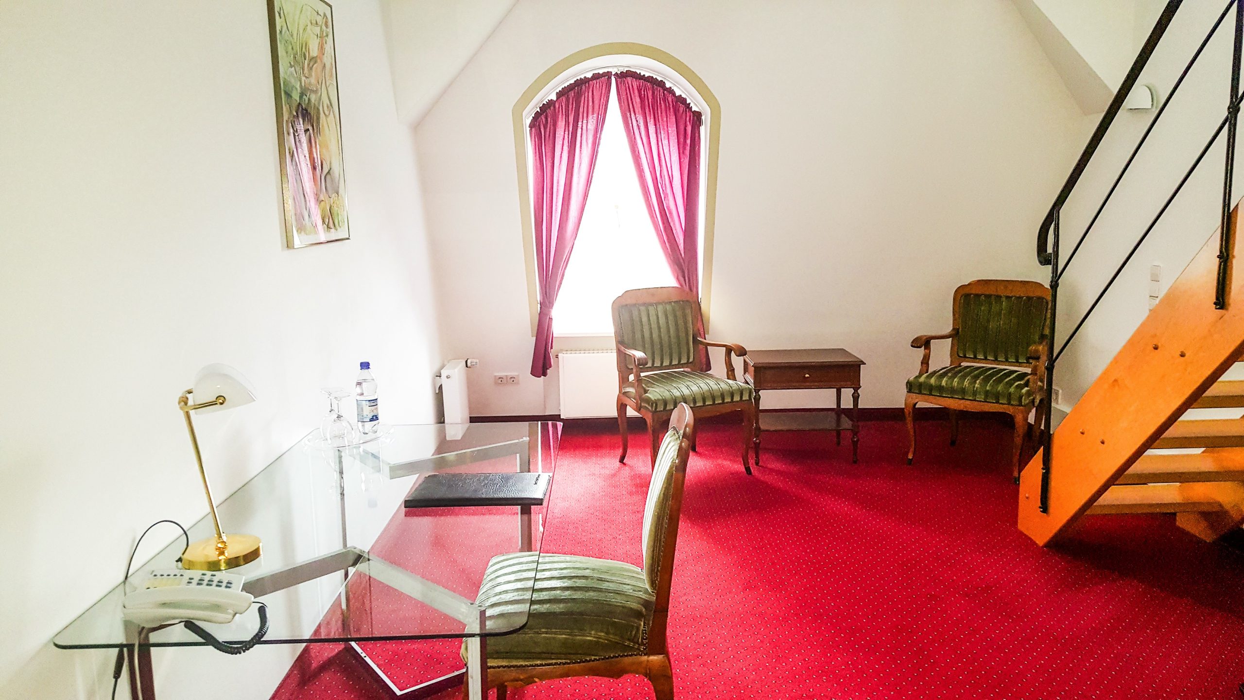 Arbeits- und Sitzbereich einer Maisonette im Hotel Schloss Storkau mit Glasschreibtisch, grünen Polsterstühlen, rotem Teppichboden und Rundbogenfenster