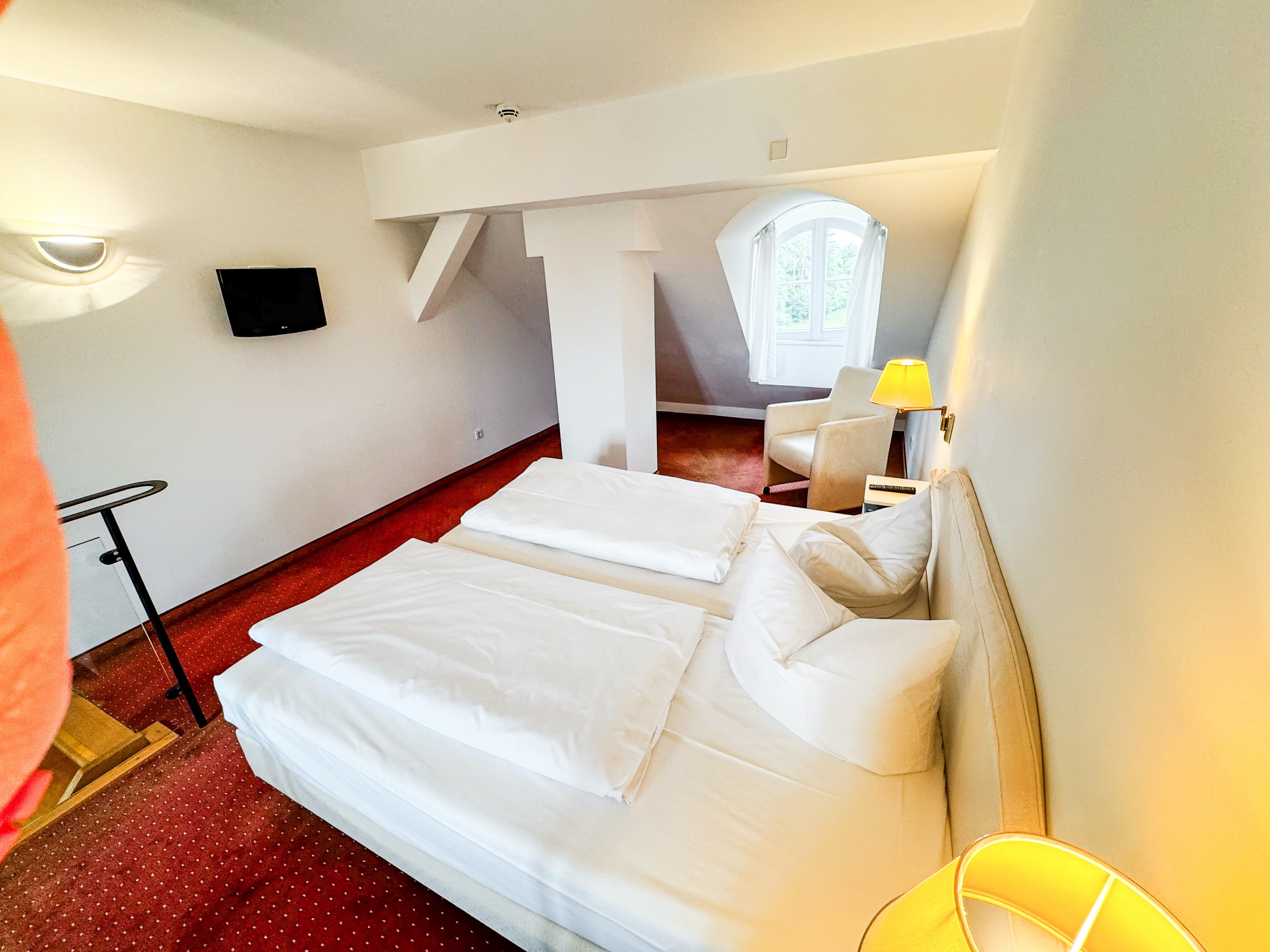 Schlafbereich unter der Dachschräge einer Maisonette im Hotel Schloss Storkau mit Doppelbett, Wand-TV und Sessel am Bogenfenster