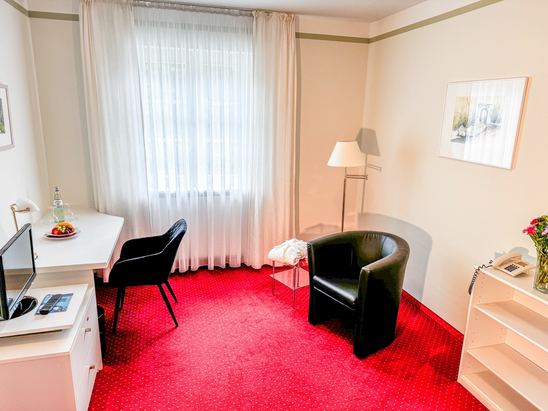 Standard-Zimmer im Hotel Schloss Storkau mit weißem Schreibtisch, schwarzem Sessel, Stehlampe und rotem Teppichboden