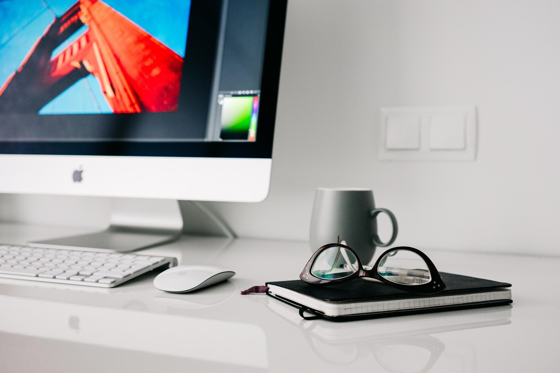 Arbeitsplatz mit Computer Aufgeräumter Schreibtisch mit iMac, Tastatur, Maus, grauer Tasse und Brille auf einem Notizbuch