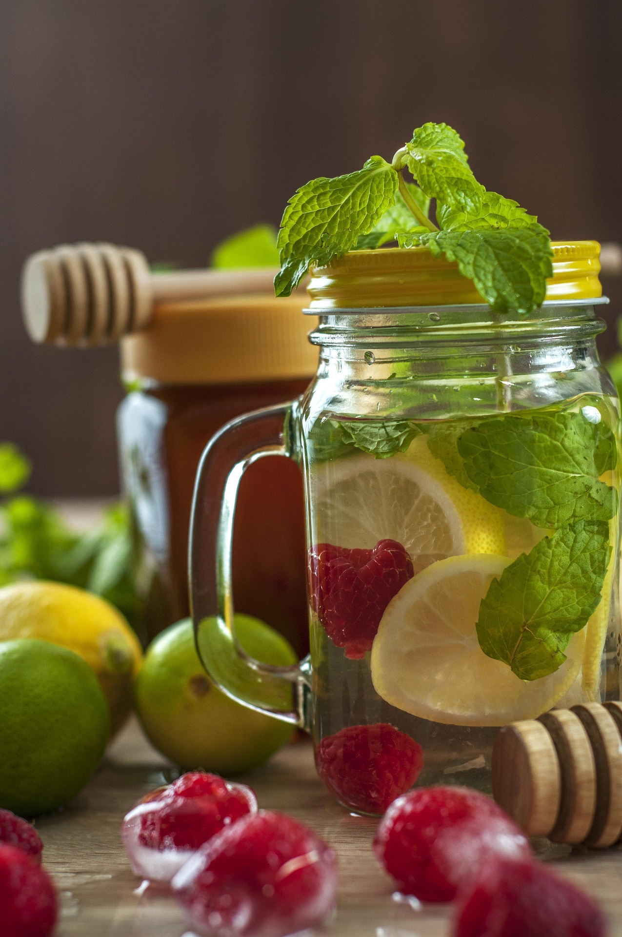 Erfrischungsgetränk mit Zitrone und Minze Henkelglas mit Wasser, Zitronenscheiben, Minze und Himbeeren, dahinter ein Honigglas mit Honigheber