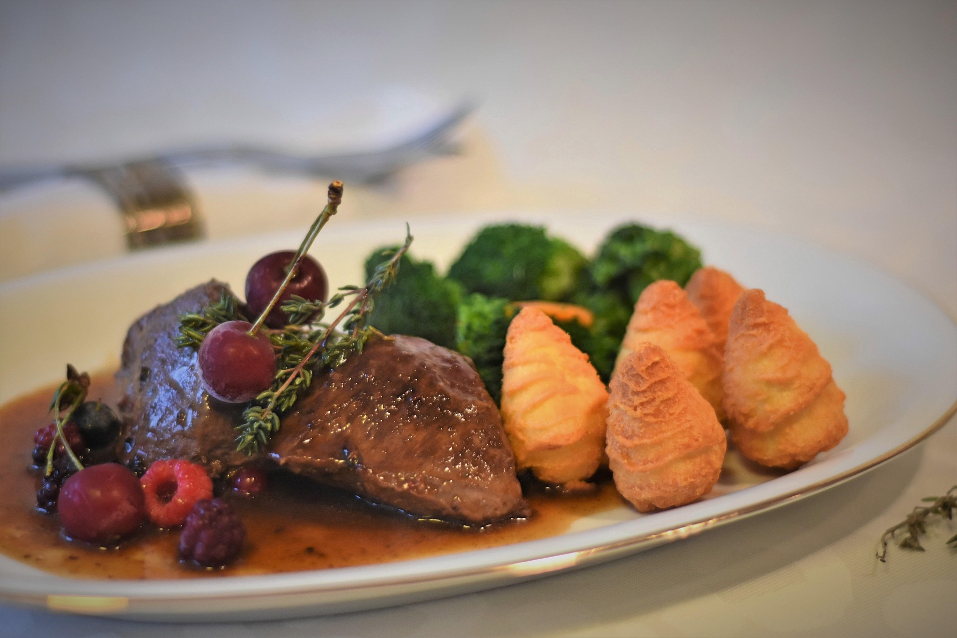 Fleischgericht mit Herzoginkartoffeln Geschmorte Fleischmedaillons in dunkler Sauce mit Herzoginkartoffeln, Brokkoli und frischen Beeren auf weißem Teller