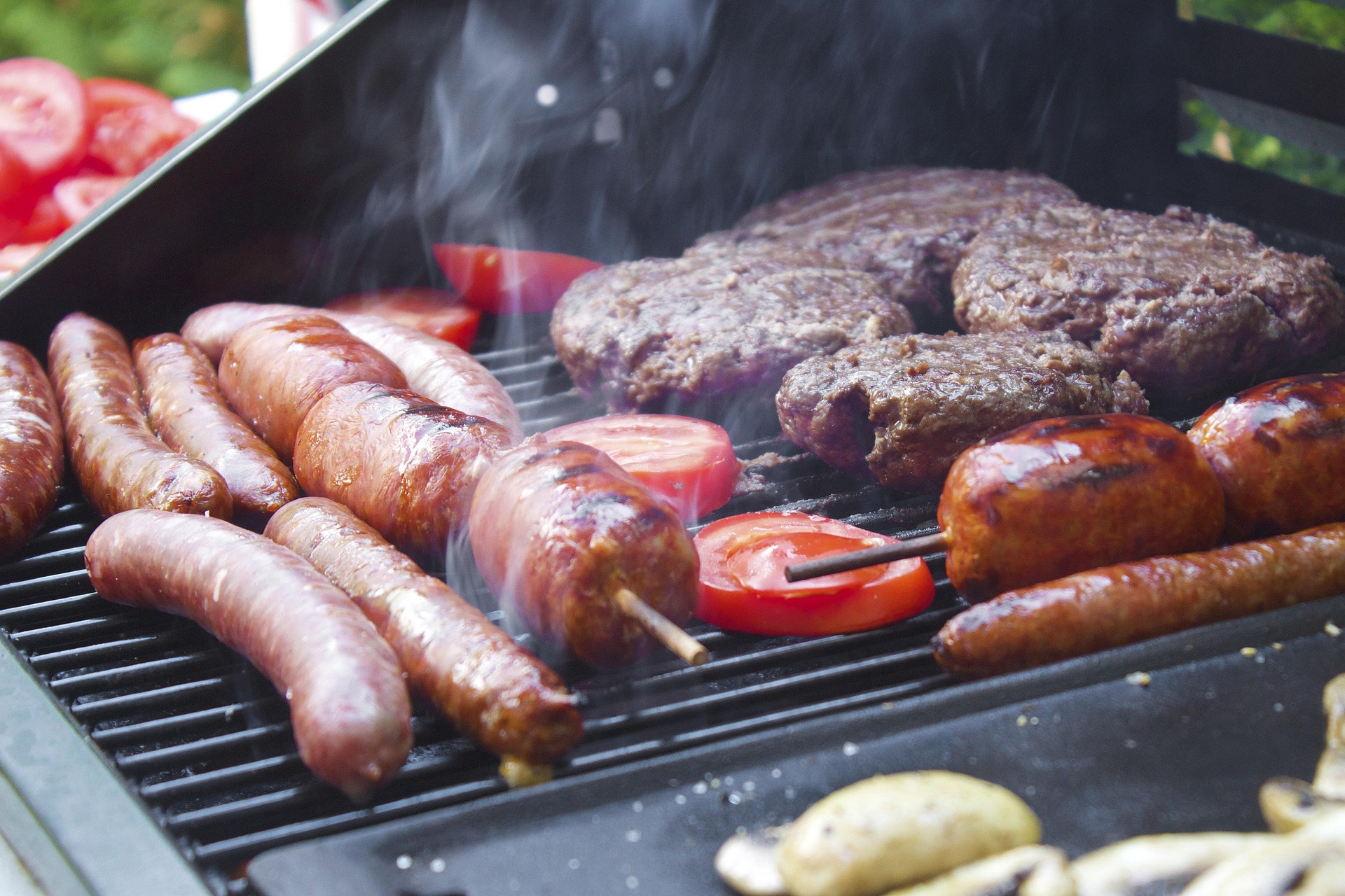 Gegrilltes auf dem Rost Würstchen, Frikadellen und Tomatenscheiben brutzeln auf einem rauchenden Grillrost
