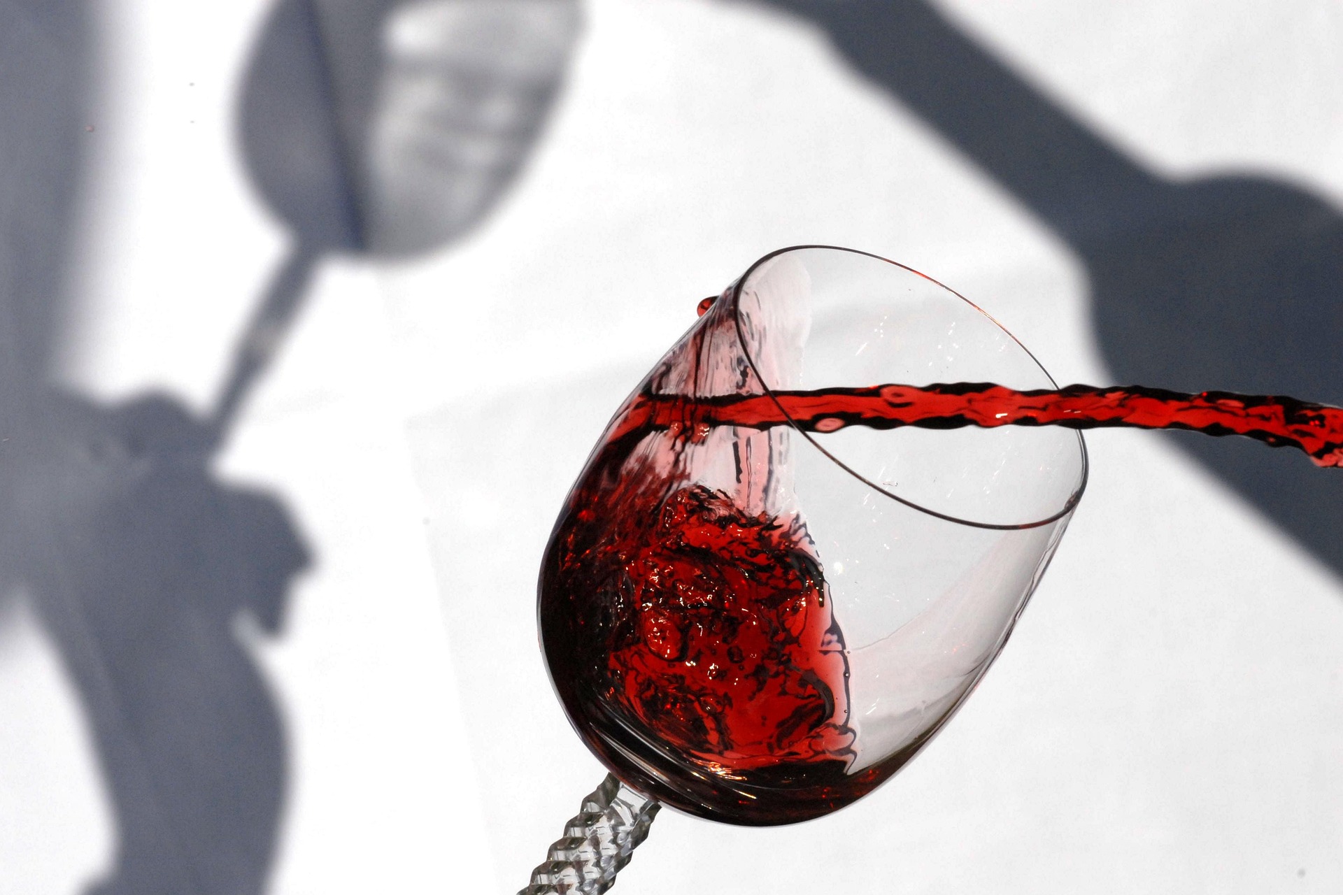 Rotwein wird in ein geneigtes Weinglas eingeschenkt, im Hintergrund Schattenspiel auf weißem Grund