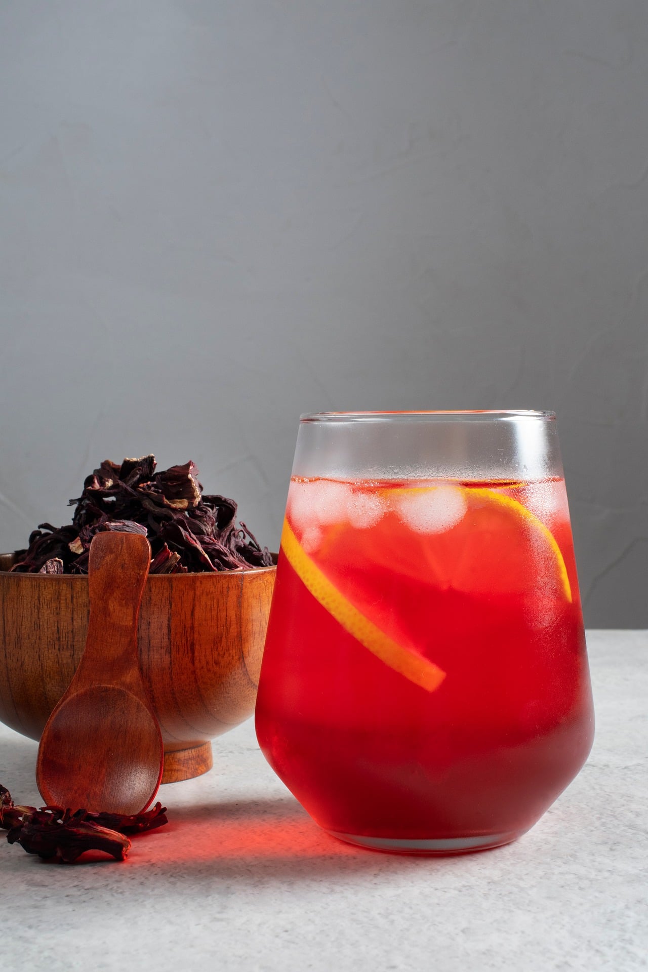 Hibiskustee mit Zitrone Glas mit rotem Hibiskus-Eistee, Eiswürfeln und Zitronenscheibe neben einer Holzschale mit getrockneten Hibiskusblüten